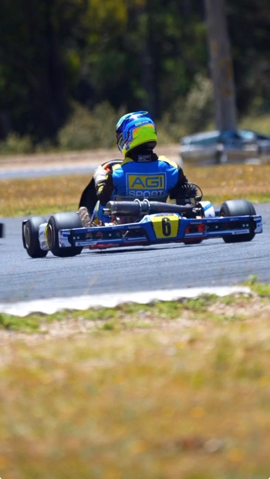 A go-kart driver in a racing suit and helmet driving a blue and yellow go-kart with the number 6, on an outdoor track.