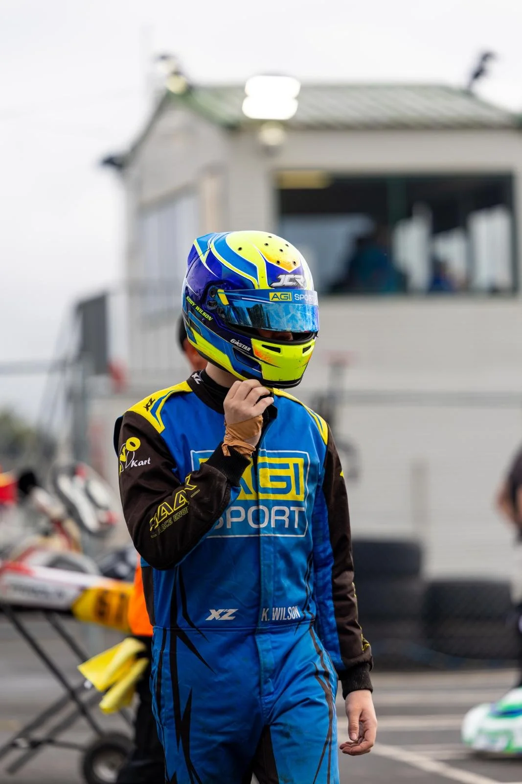 A person in a blue and black racing suit with a yellow and blue helmet, standing on a race track, adjusting their helmet. There are go-karts and a building in the background.