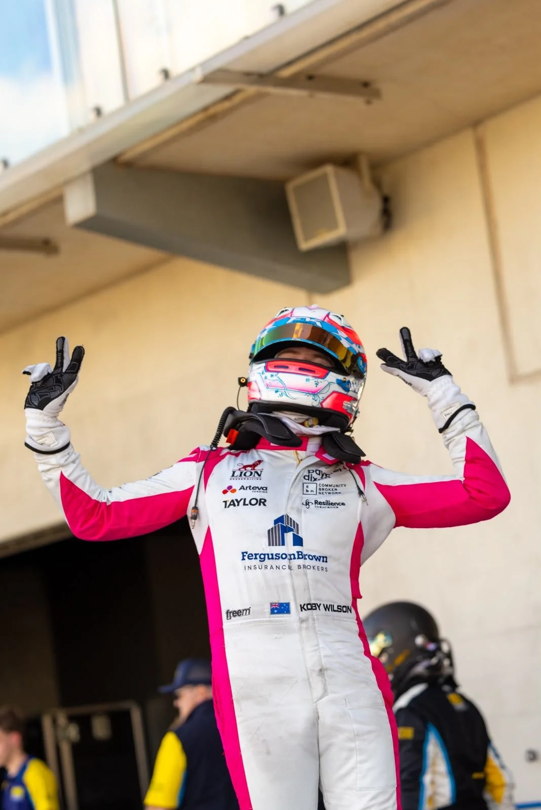 Race car driver in a white and pink racing suit and helmet making victory signs with both hands.