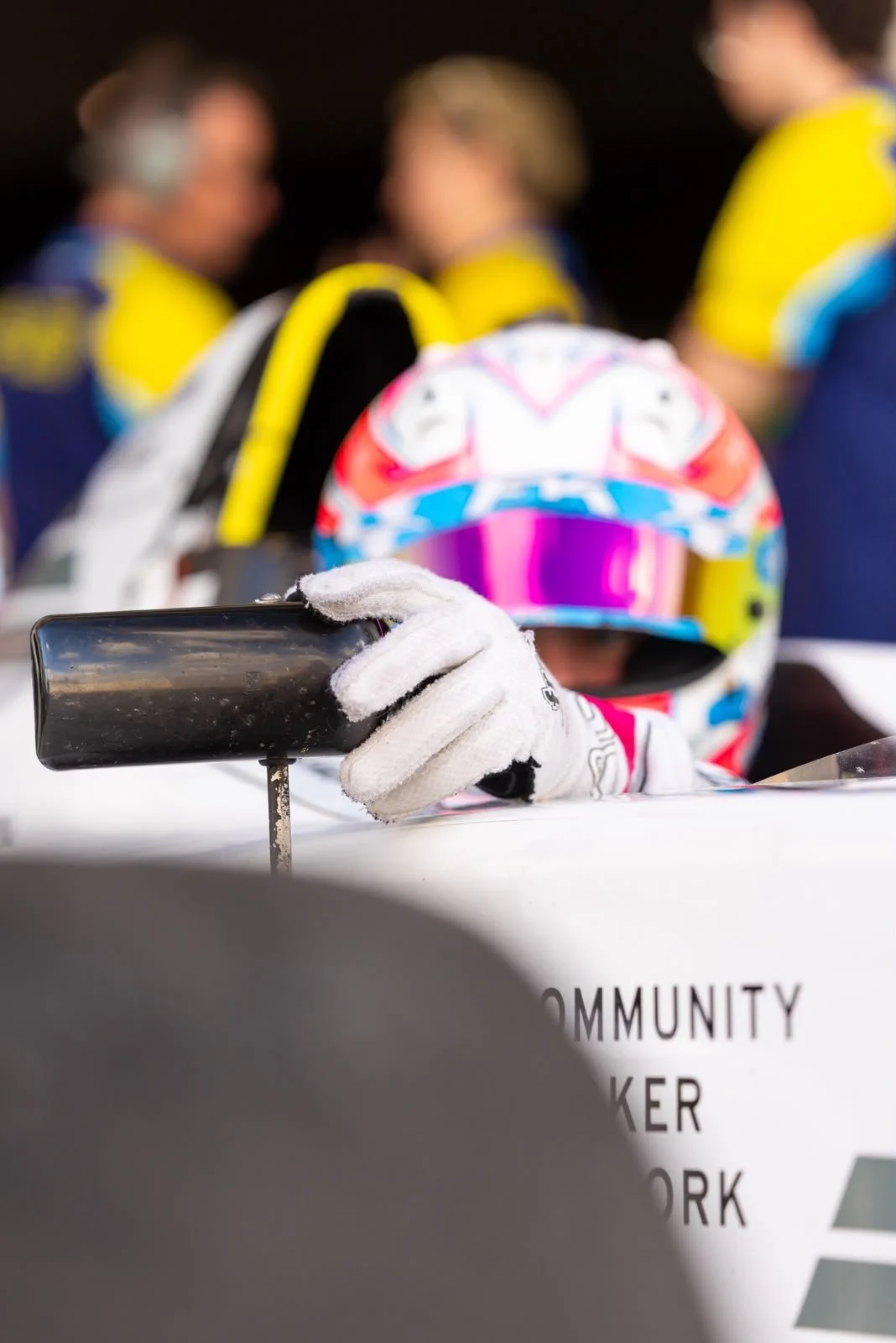 Person in colorful helmet and white gloves holding a camera or phone, with a blurred background of people wearing yellow and blue shirts and a partially visible sign reading 'COMMUNITY'.