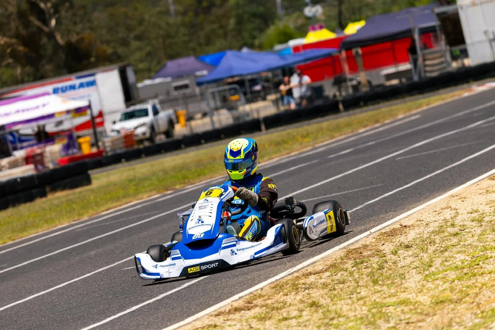 A person racing a go-kart on a race track, wearing a colorful helmet and racing suit, with tents and vehicles visible in the background.