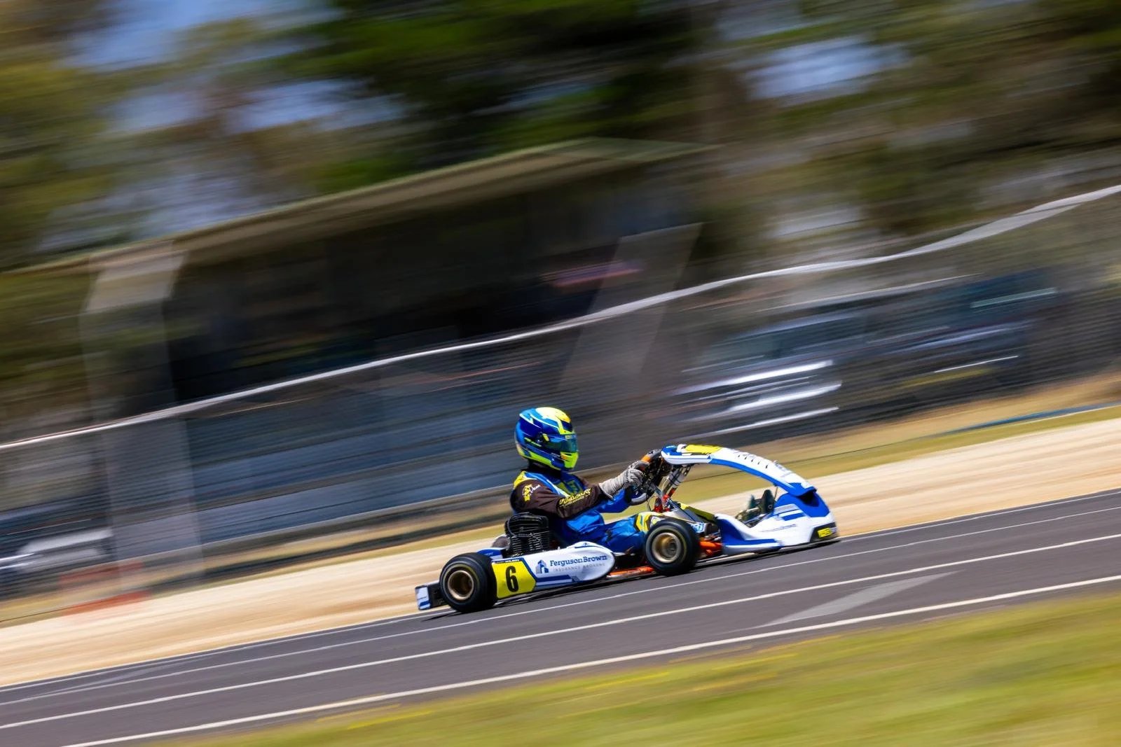 A person racing in a go-kart on a track during daytime, wearing a racing suit and helmet, with background motion blur indicating high speed.