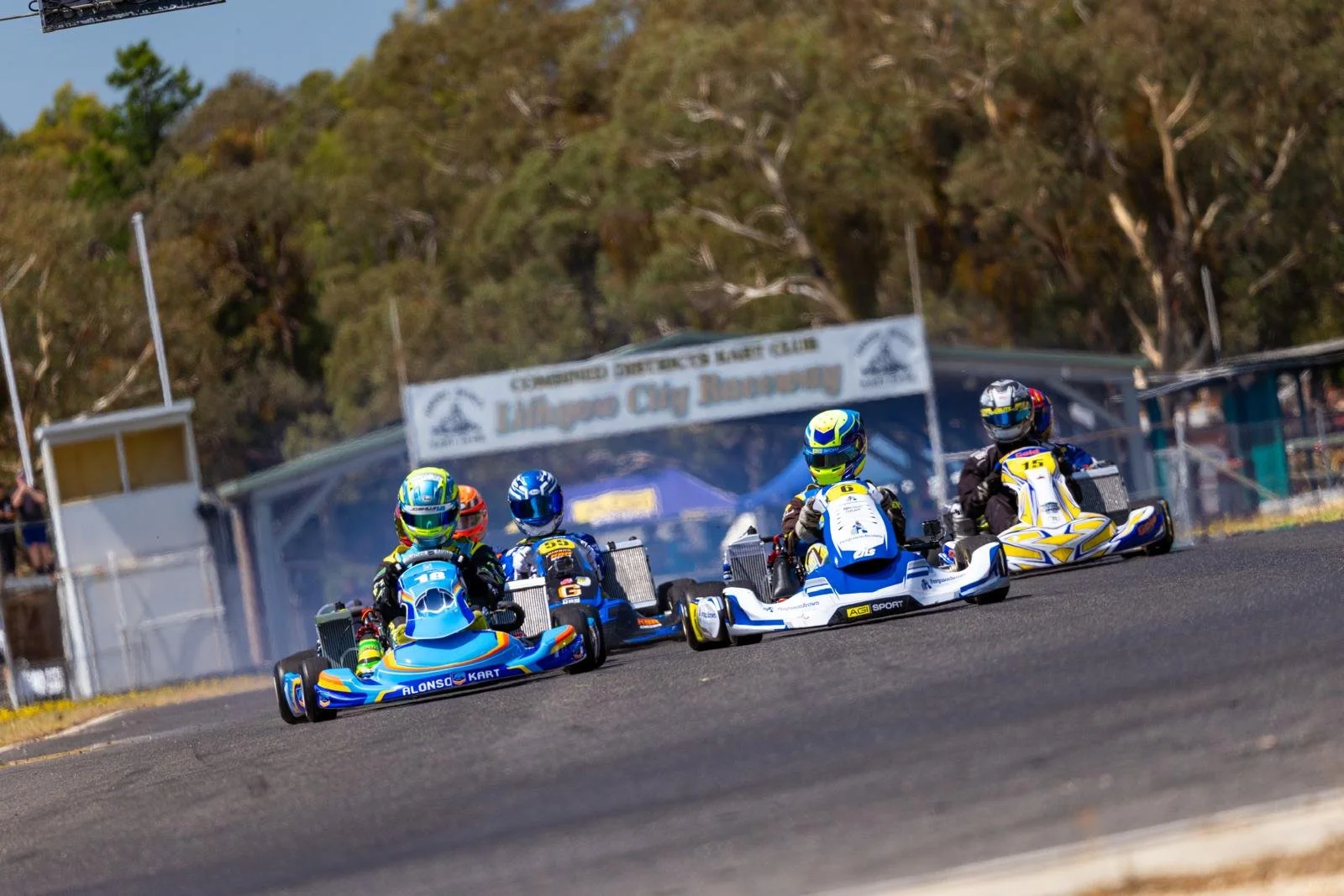 Four go-karts racing on a track with a sign that reads 'Lillico City Raceway' in the background, surrounded by trees.