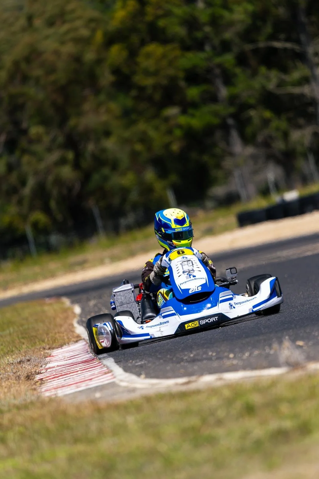 A person racing in a go-kart on a track, wearing a helmet and racing gear, with trees and tires visible in the background.