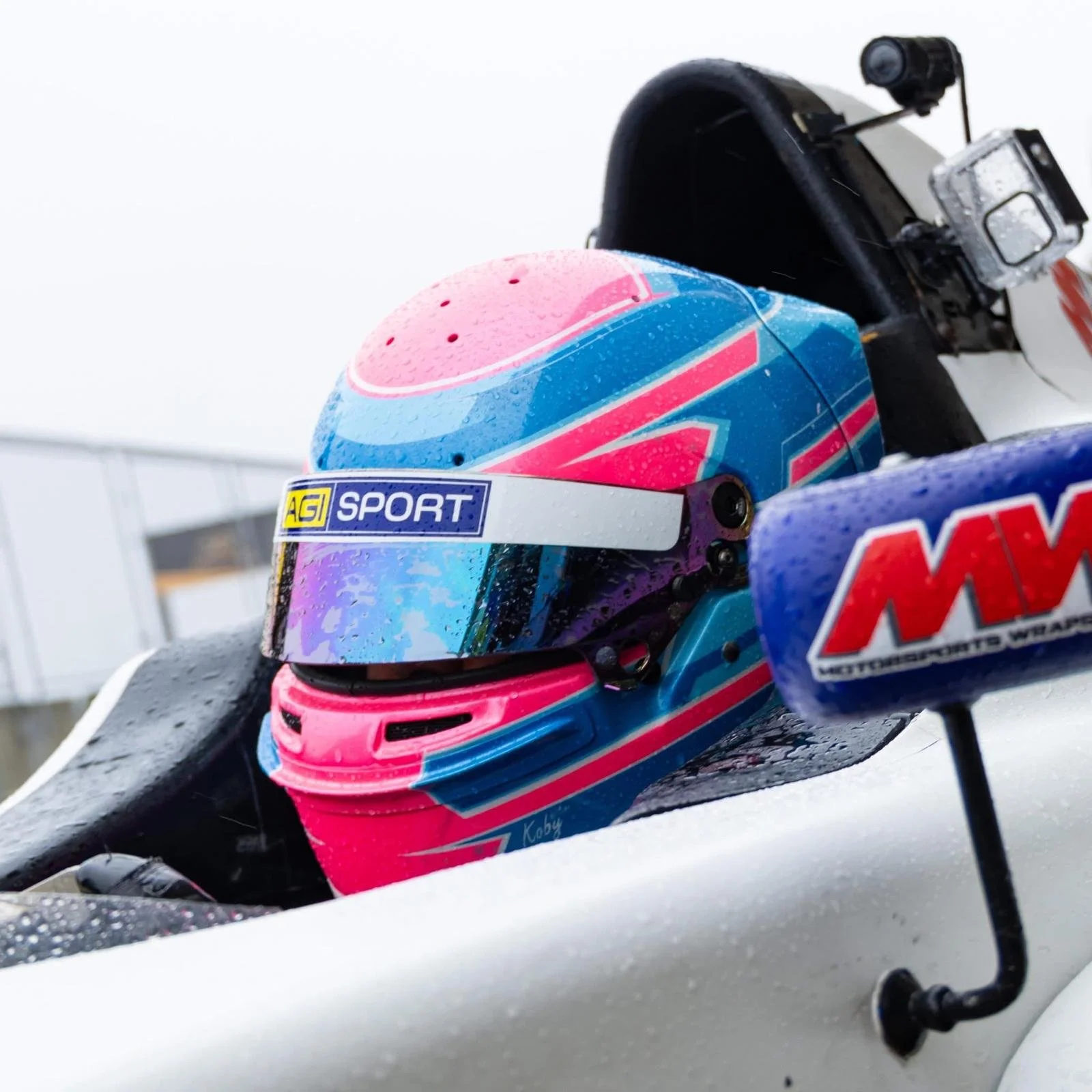 A racing helmet with pink, blue, purple, and teal graphics, resting on a wet surface next to a racing vehicle with a sponsorship sticker. The helmet has a reflective visor and droplets of rain on it.