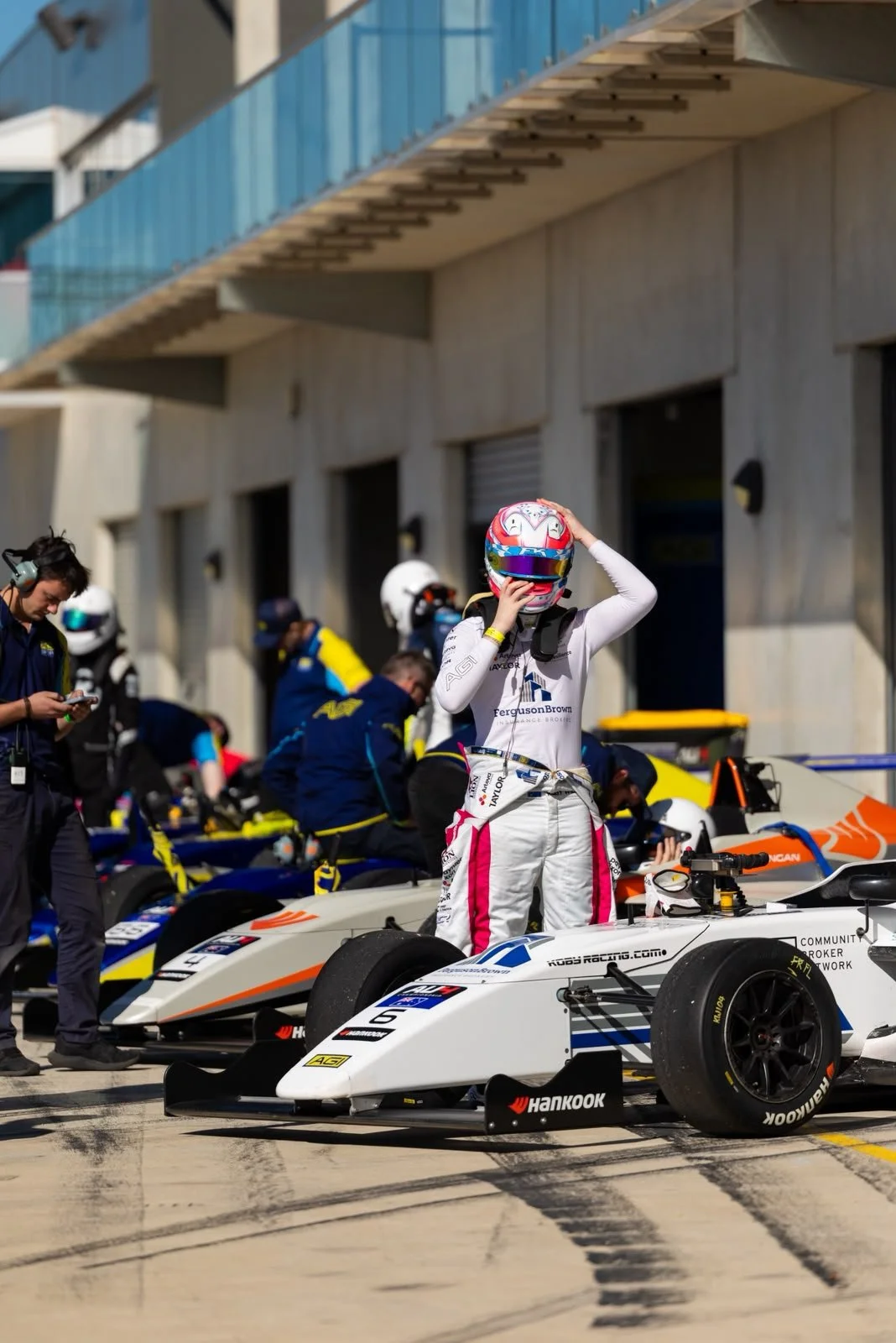 A race car driver in a white racing suit adjusts their colorful helmet while standing beside a white Formula racing car in a pit area, with team members and other cars in the background.
