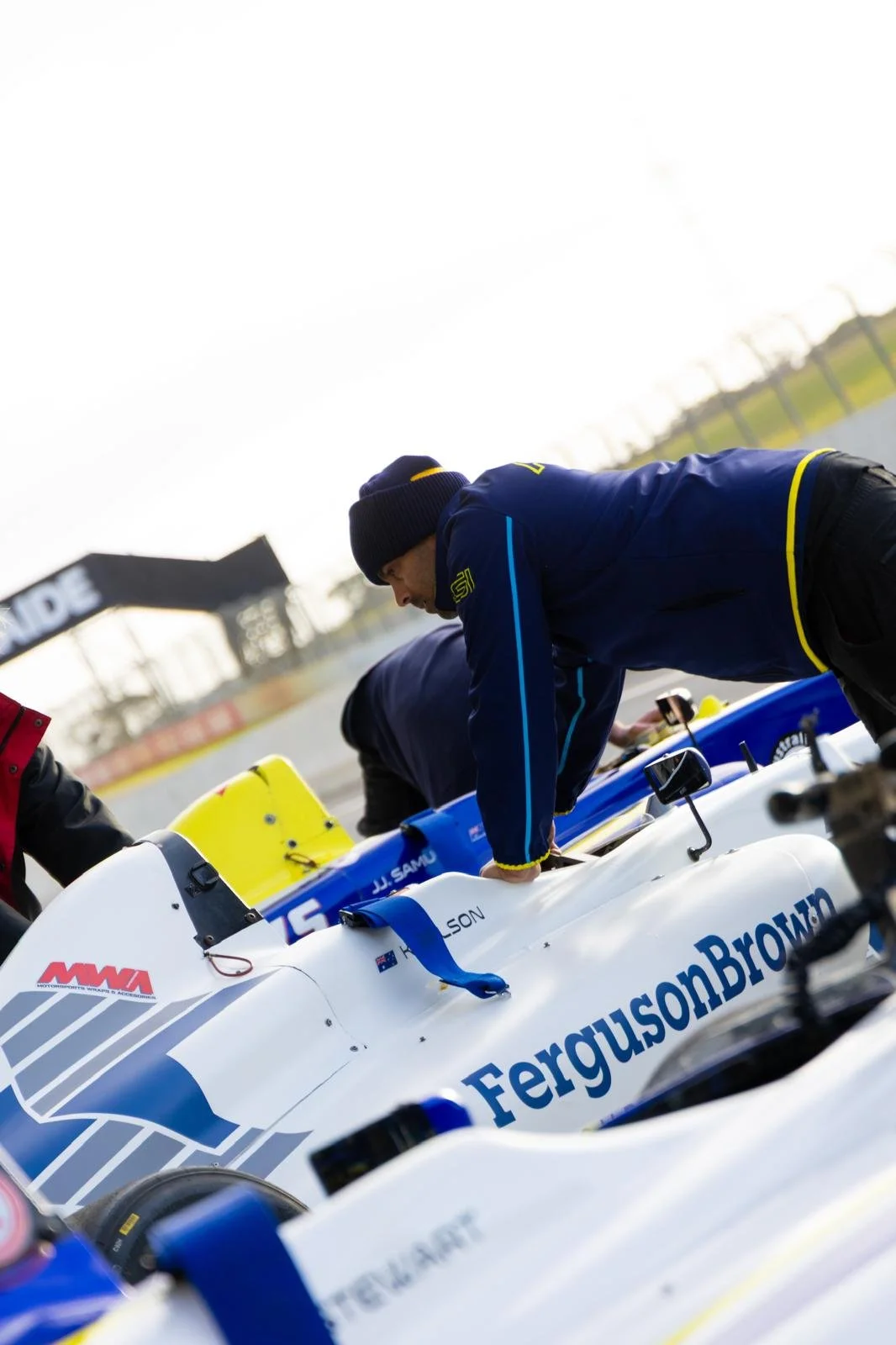 A person in a blue jacket and black beanie preparing a race car on a race track, with another car in the background and a starting line in the distance.