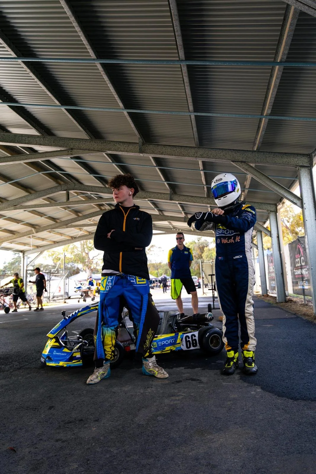 Two race car drivers in racing suits and helmets stand under a metal shelter with a go-kart between them. Several people are in the background at a racing event.