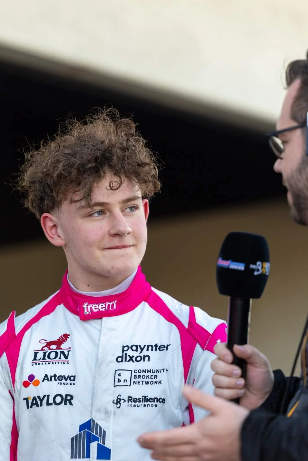 Young male race car driver with curly hair being interviewed, holding a microphone, wearing a racing suit with various sponsor logos.