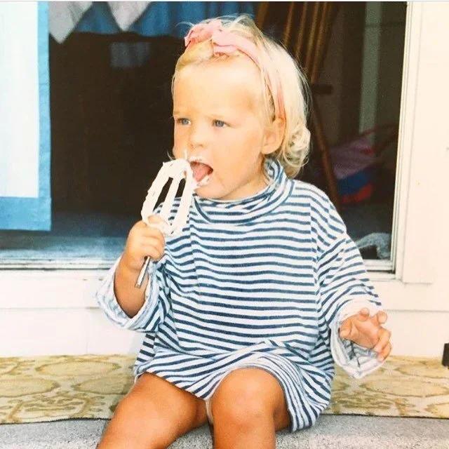 A young girl with blonde hair and a pink headband sitting on the floor, licking a spatula with white frosting, wearing a blue and white striped dress.