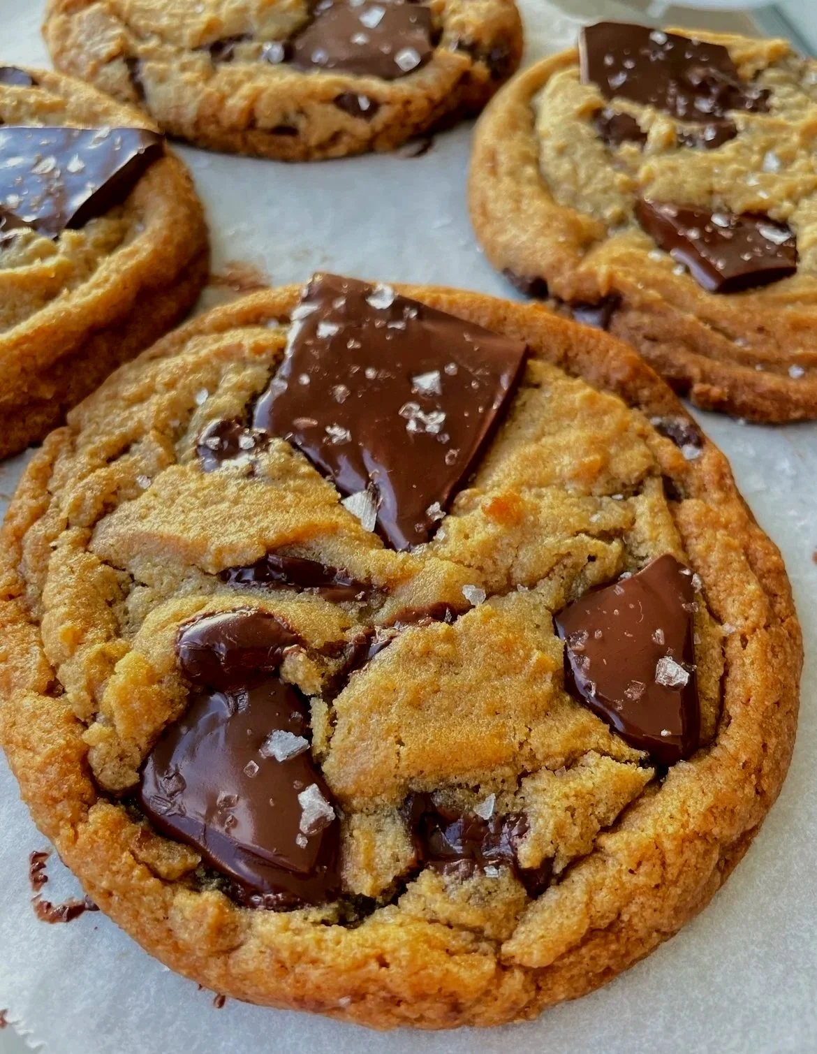 Close-up of four chocolate chip cookies with large chocolate chunks and a sprinkle of sea salt on top, resting on a piece of parchment paper.
