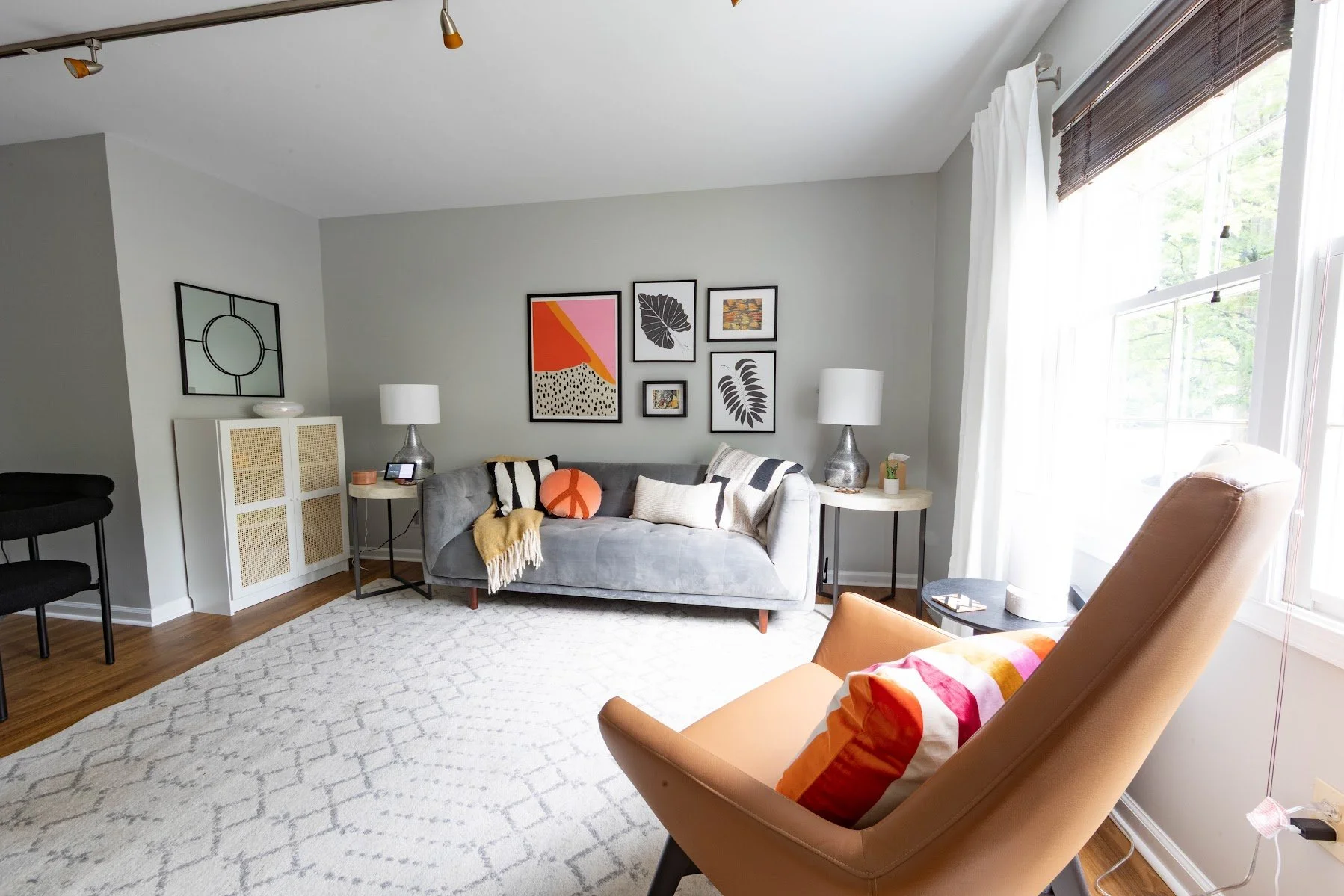 Living room with a gray sofa, black and white pillows, a yellow throw blanket, and abstract wall art. Features a tan armchair with a colorful pillow, side tables with lamps, a white cabinet, and large windows with white curtains.