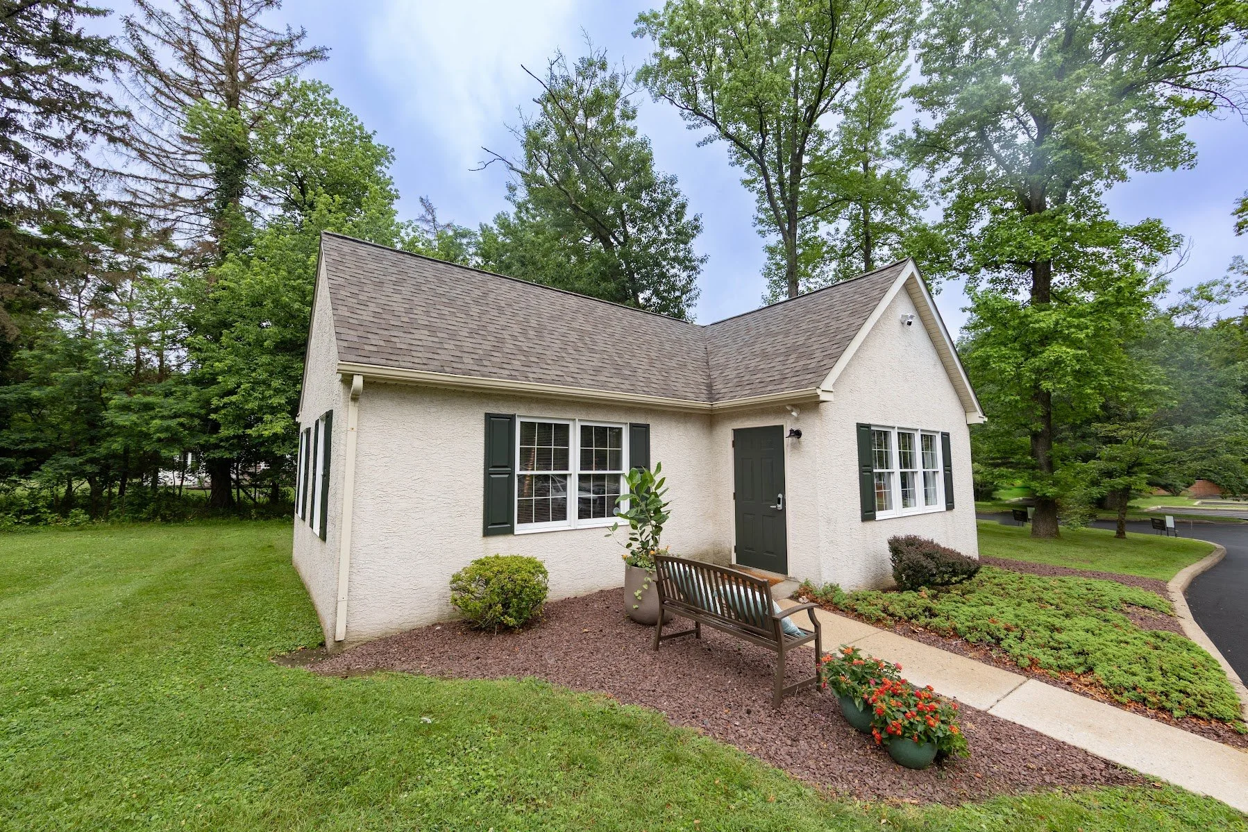 A white house with dark green shutters and a black front door, surrounded by a well-maintained yard with bushes, potted plants, and a bench, with tall trees in the background.