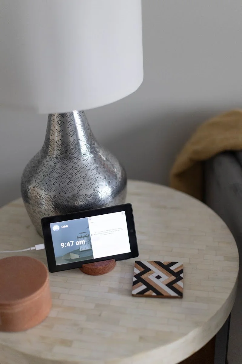 A bedside table with a modern lamp, a smartphone displaying the time 9:47 am and a weather app, a small round container, and a decorative coaster with a geometric pattern.