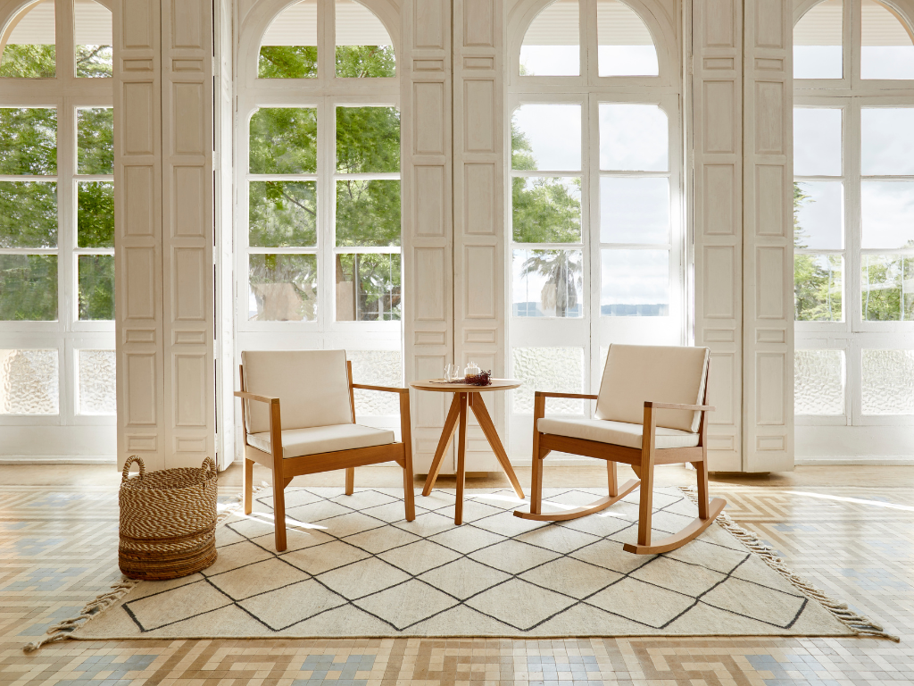 A bright sunlit room with large windows, two wooden chairs with white cushions, a small wooden table with a decorative item, a woven basket, and a patterned rug on a tiled floor.