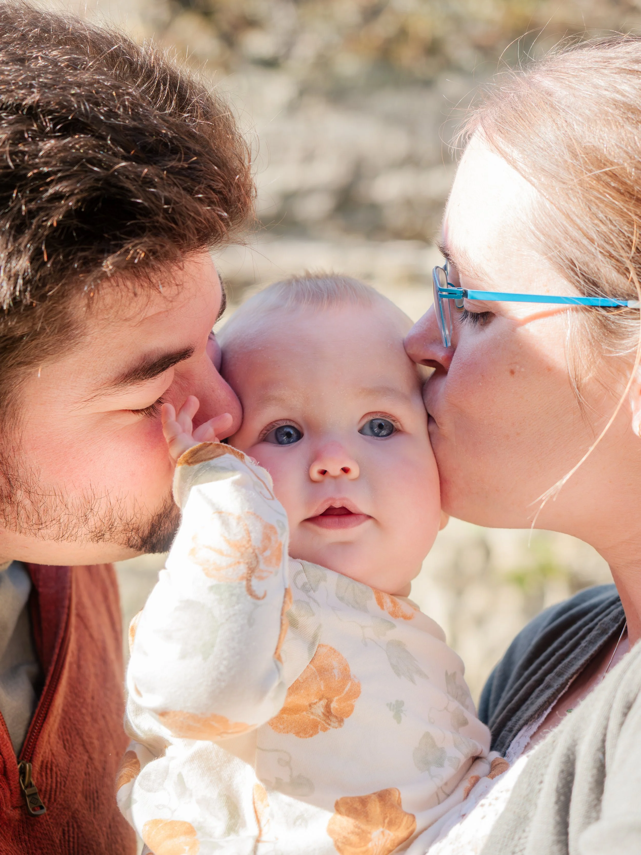 A young man and woman kissing their baby on each cheek outdoors in sunny weather.