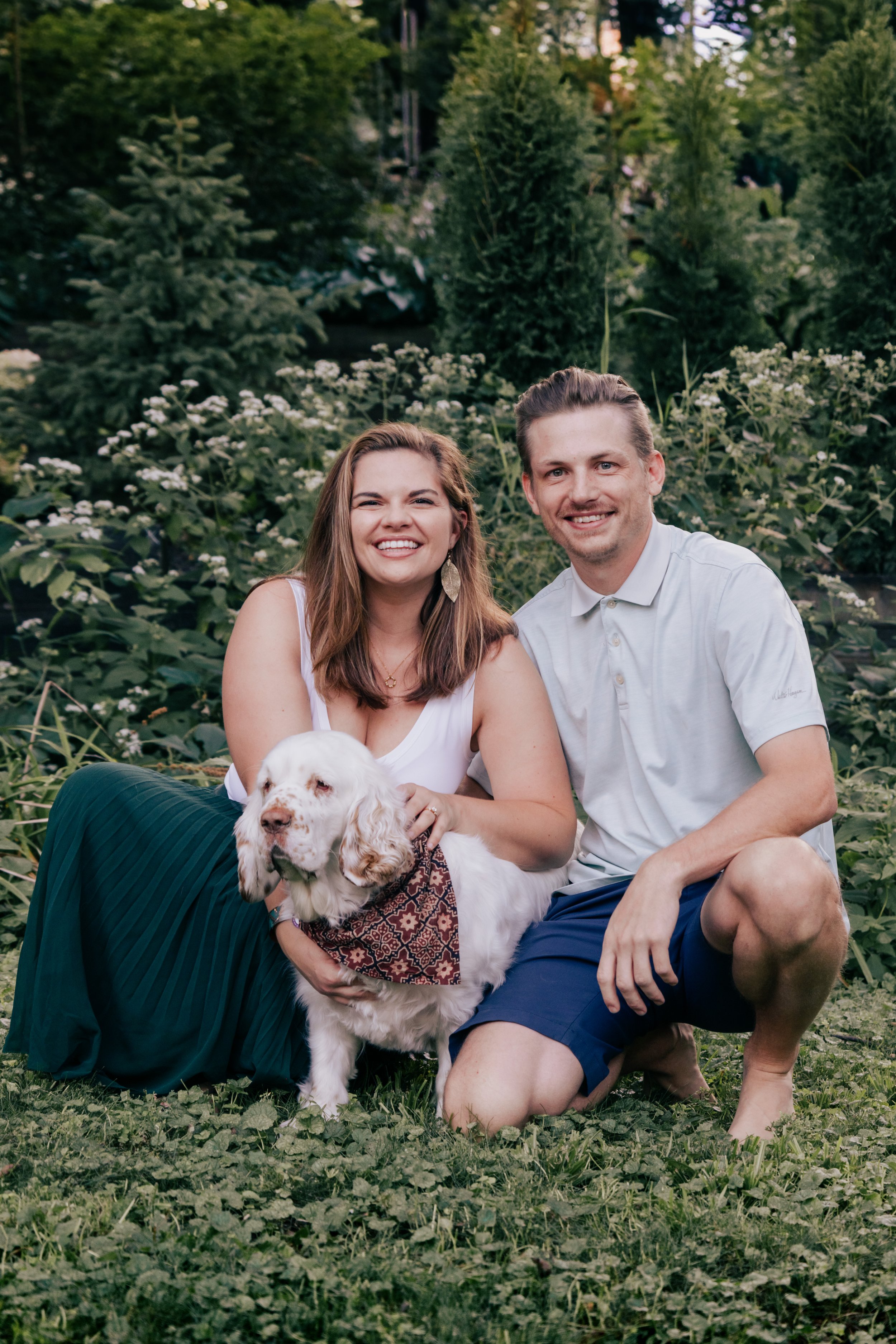 An engagement portrait of a smiling young woman and young man sitting on grass outdoors with a white puppy, surrounded by greenery.