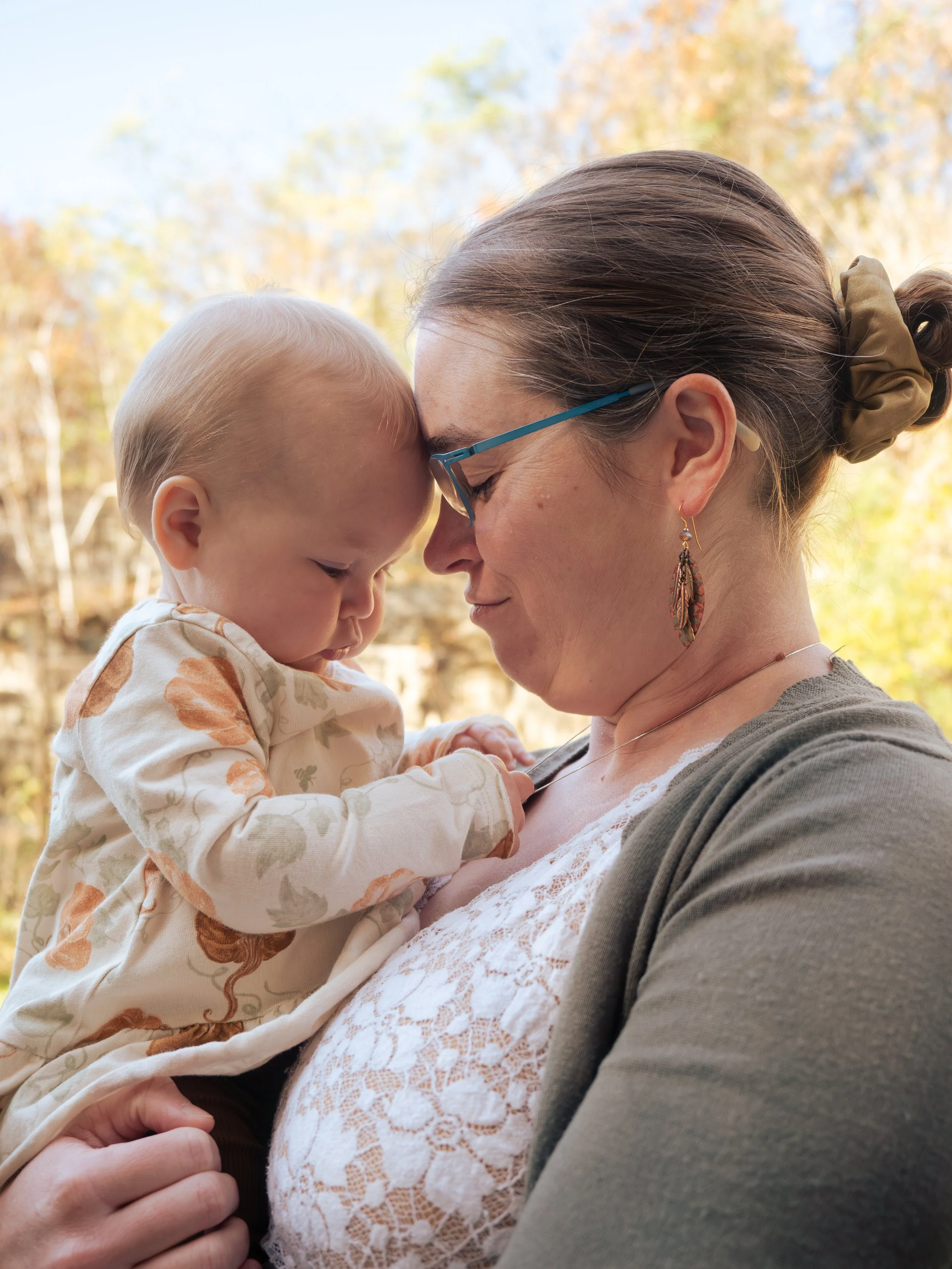 A woman holding a young child close with their foreheads touching outdoors during fall.