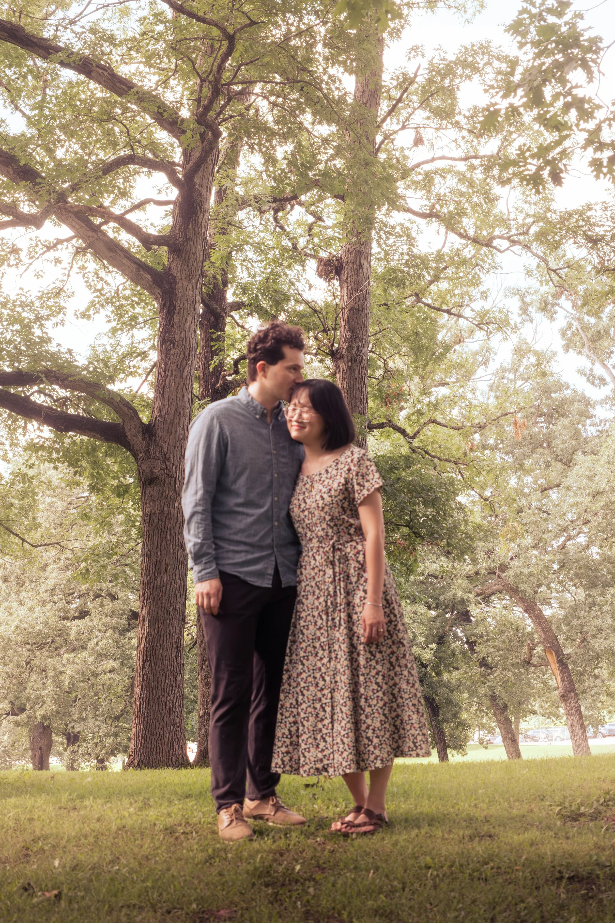 A couple standing closely together in a park with tall trees and green grass, the man whispering into the woman's ear.