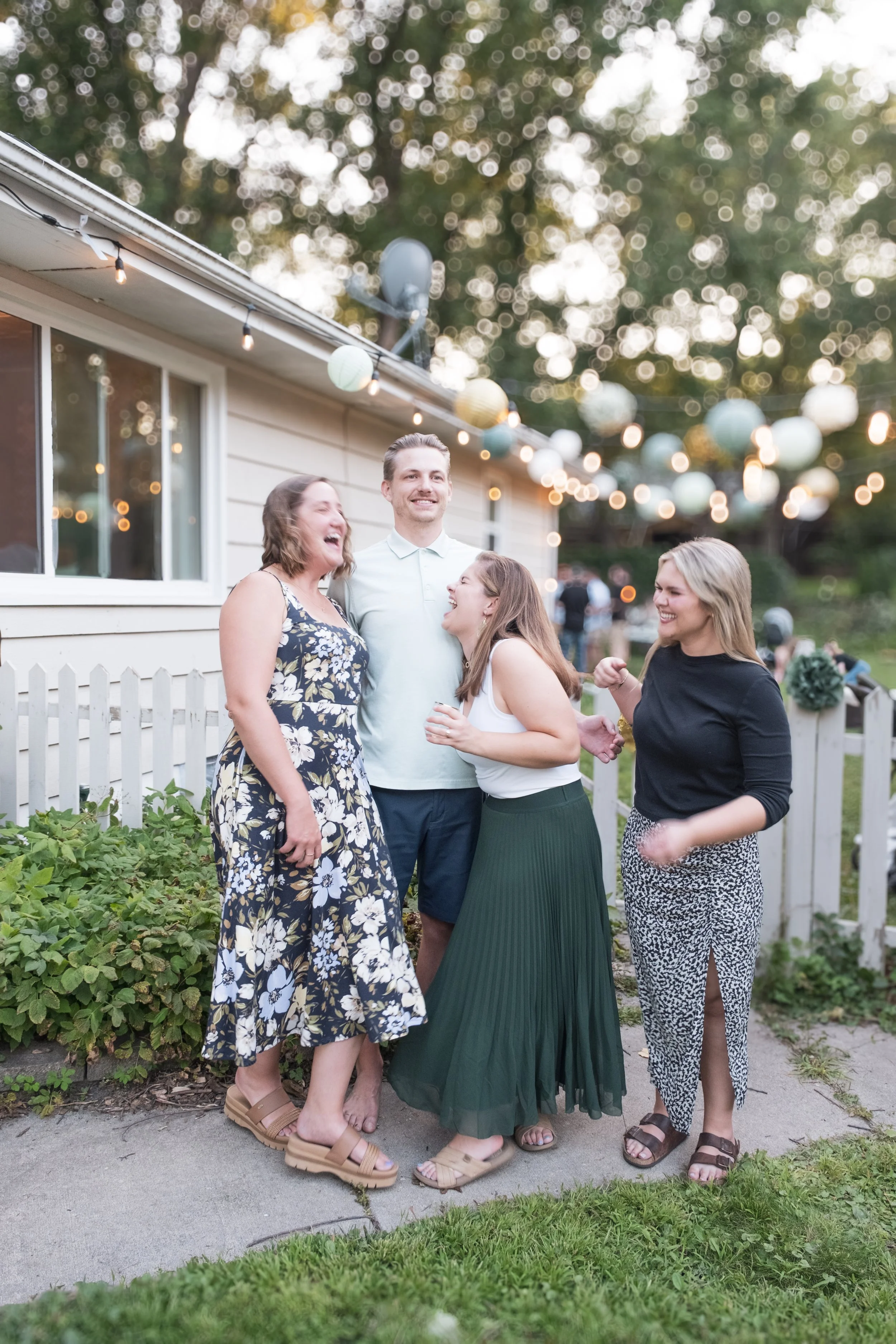 Four friends laughing and talking outdoors during an evening gathering in a backyard decorated with string lights and paper lanterns.