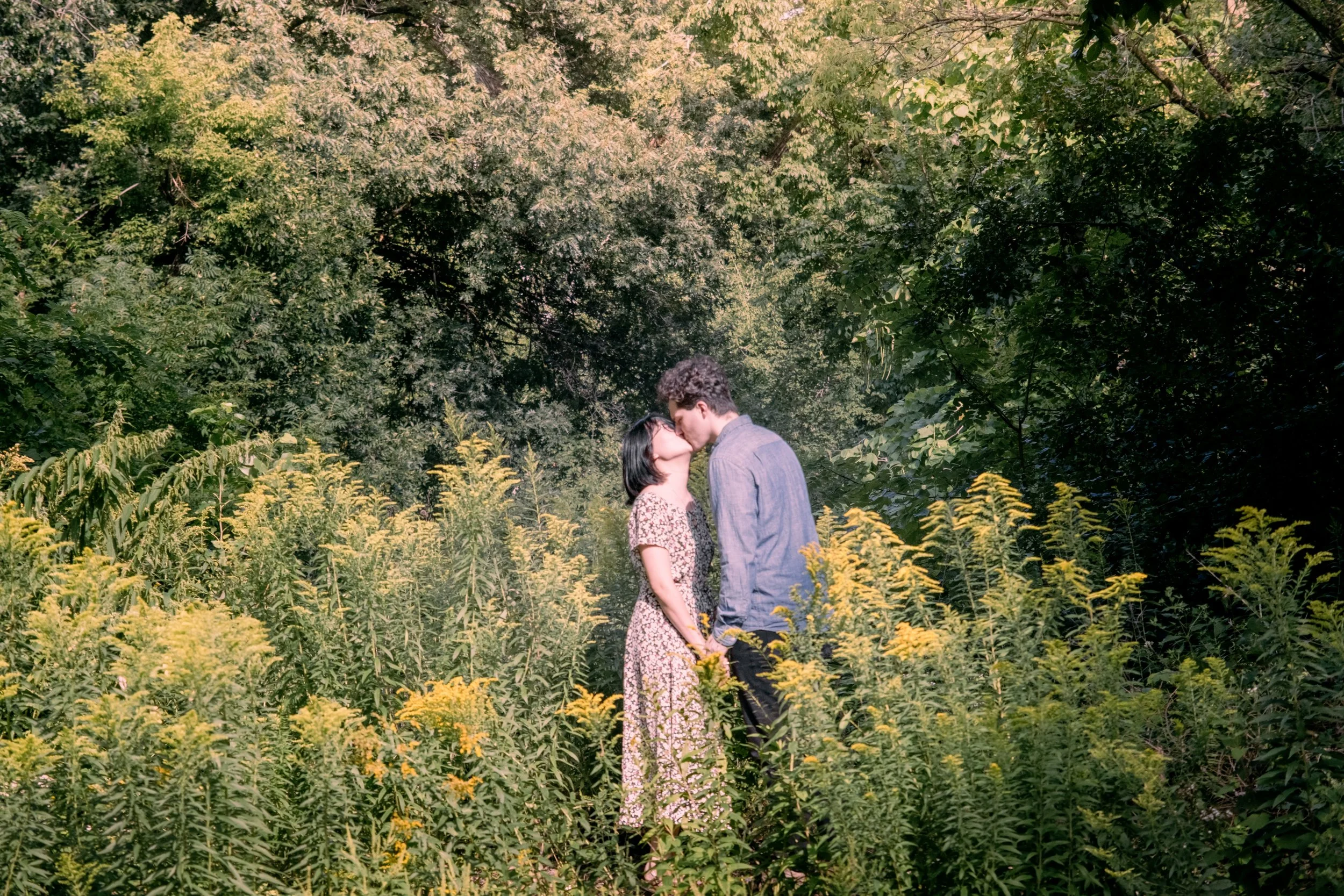 A couple kissing in a lush, green outdoor setting with yellow wildflowers and dense trees in the background.