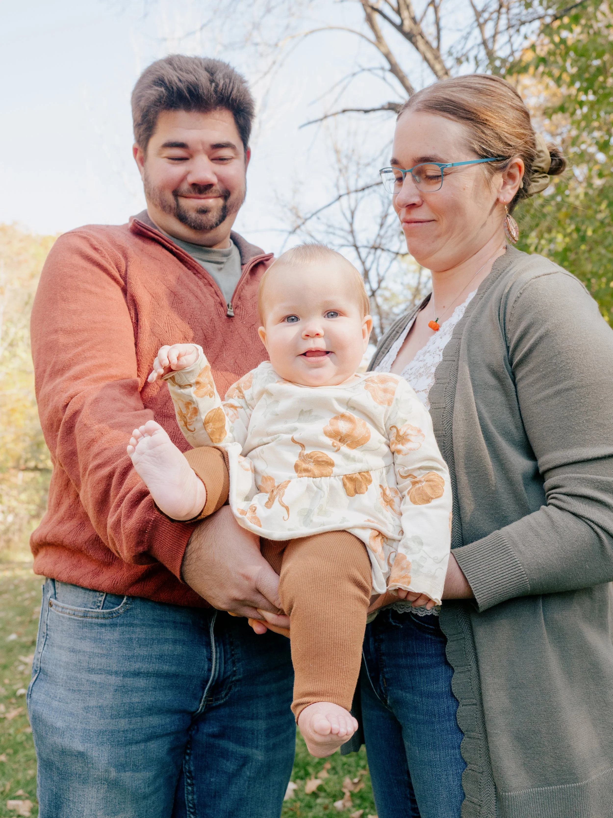 A family of three standing outdoors in autumn, with a baby girl held by the father, all smiling and dressed in casual fall clothing.