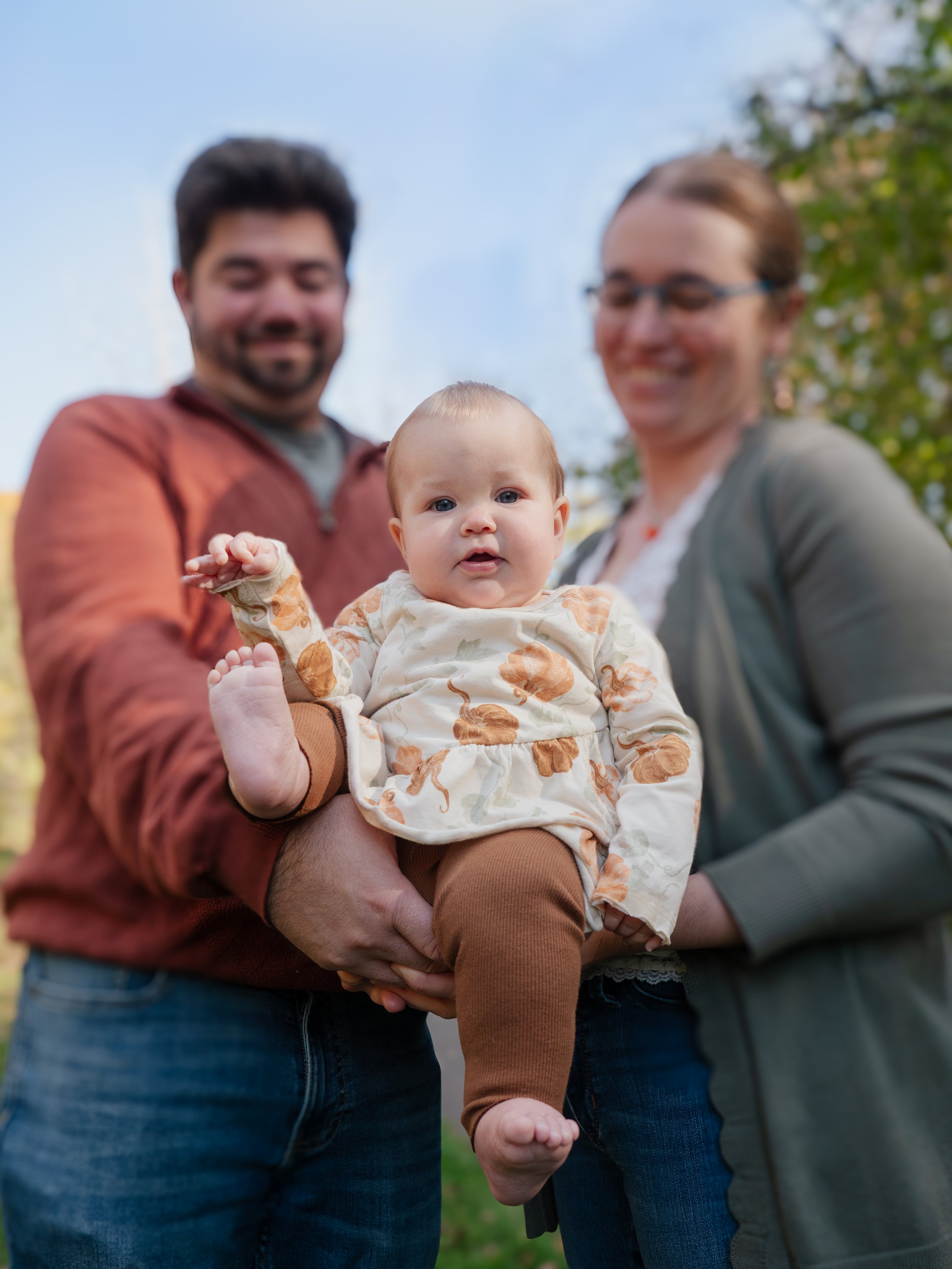 Family of three outdoors with a baby in the middle, held by two adults, with trees and a blue sky in the background.