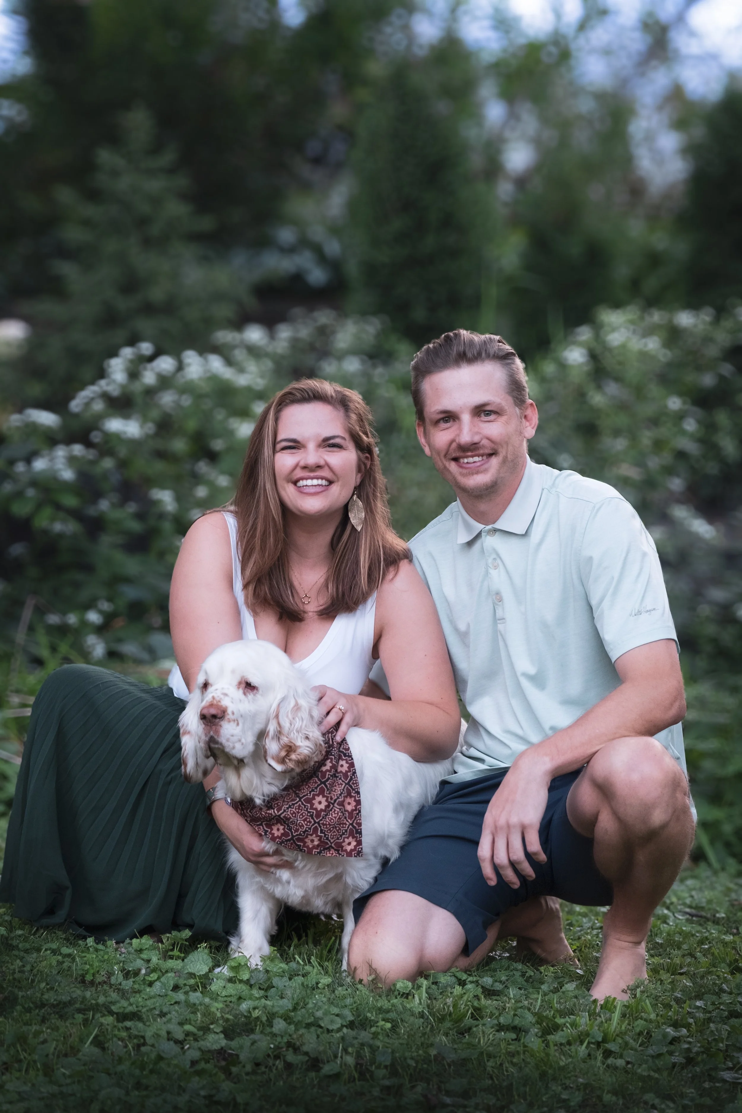 A happy couple sitting on grass with their dog outdoors. The woman has brown hair and is wearing a white sleeveless top and a long green skirt. The man has brown hair and is wearing a light-colored polo shirt and dark shorts. The dog is a white and b