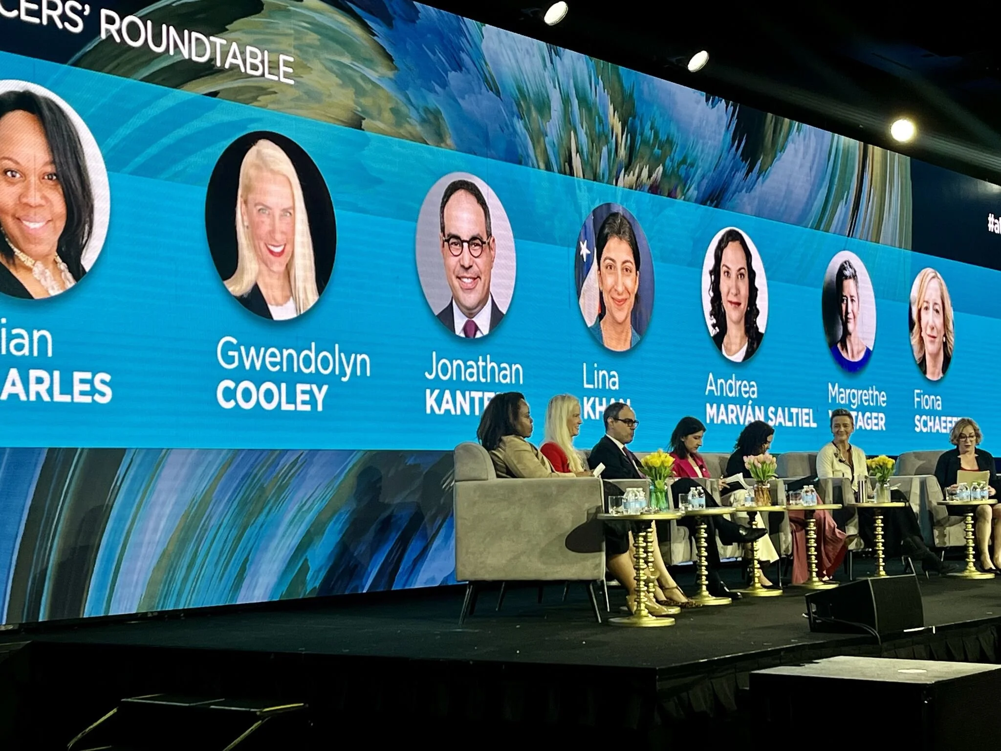 Panel discussion at a conference with seven women and two men seated on stage, with a large screen behind displaying headshots and names of speakers, including Gwendolyn Cooley, Jonathan Kanter, Lina Khan, Andrea Marván Saltiel, Margrethe Vestager.