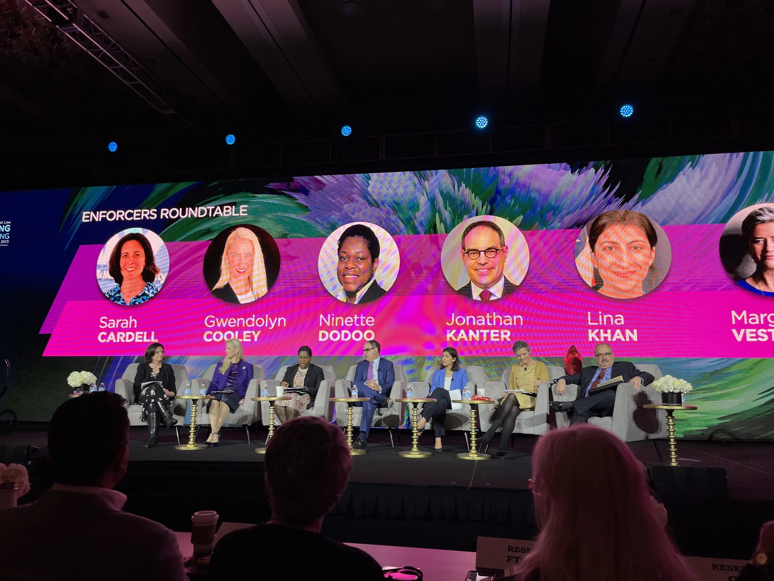 Panel discussion at a conference with seven individuals seated on stage in front of a large screen displaying their names and photos, titled 'Enforcers Roundtable'.