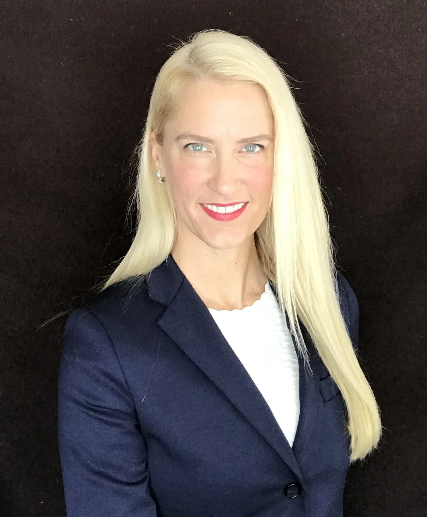 A professional woman with long blonde hair, wearing a navy blazer and red lipstick, smiling against a black background.