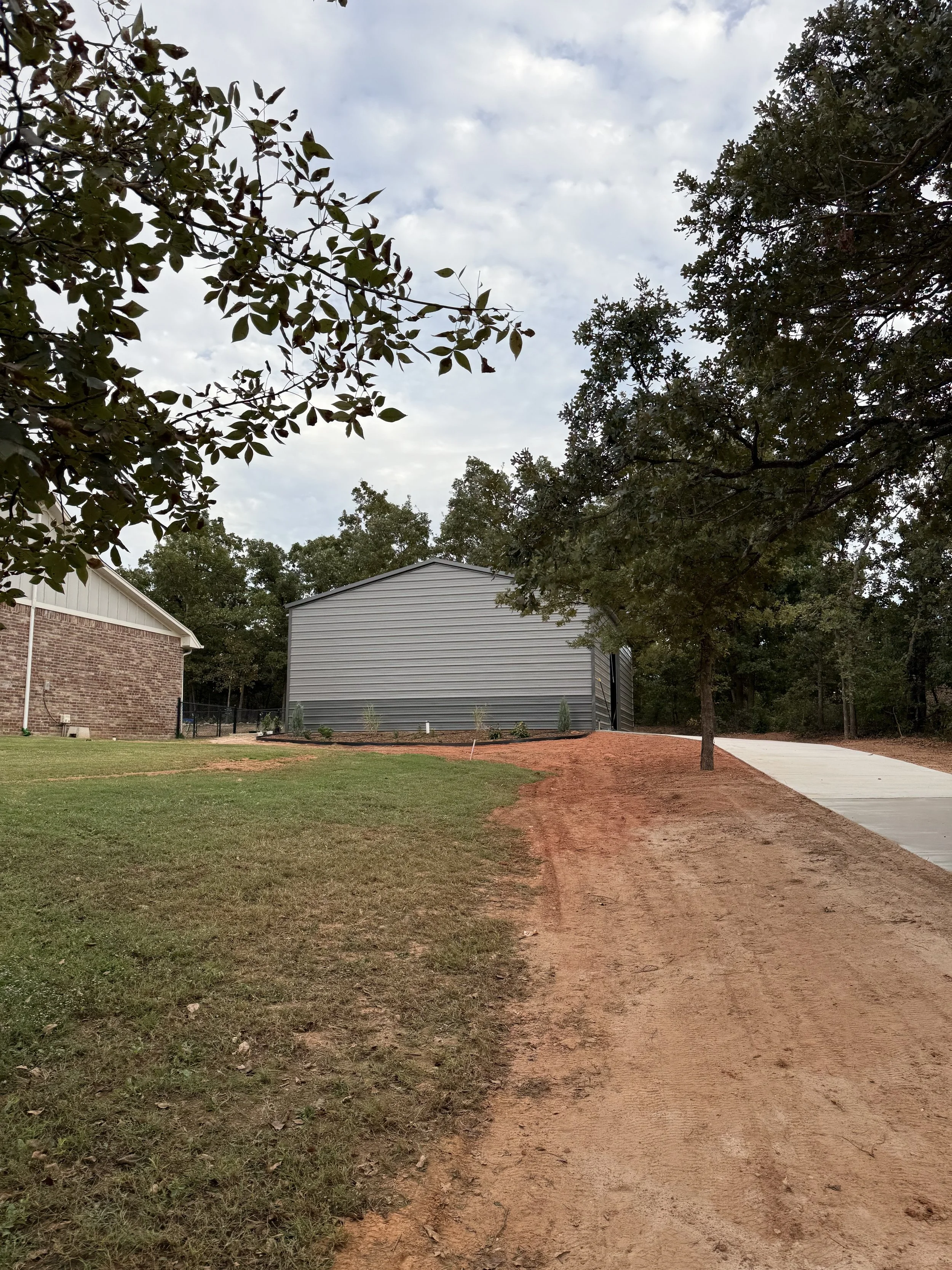 A suburban backyard with a newly paved driveway, a small tree, a grassy lawn, and a gray metal shed in the background, under a cloudy sky.