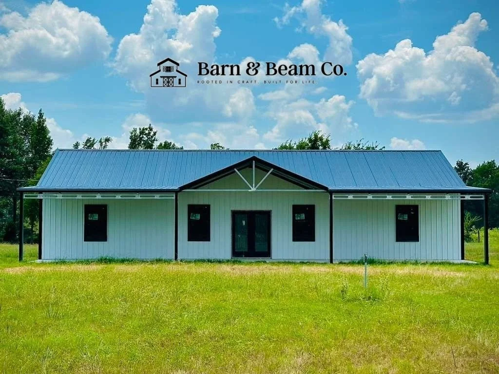 Front view of a modern barn-style building with white siding, black trim, and a blue metal roof, situated on a grassy field with trees and a blue sky with clouds in the background.