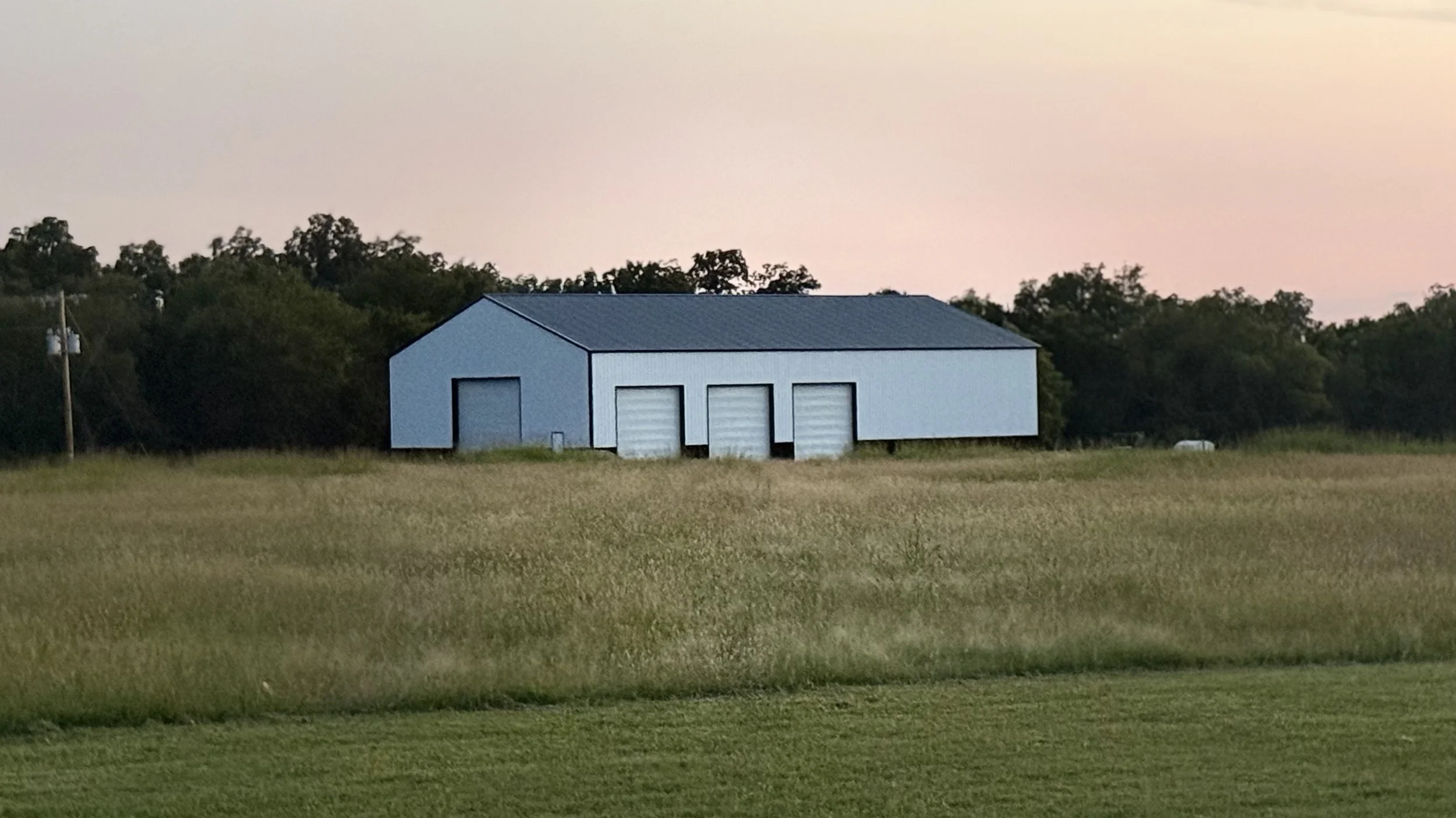 A white barn with a gray metal roof and three large white garage doors situated in a grassy field during sunset. Trees are in the background with a pinkish sky.