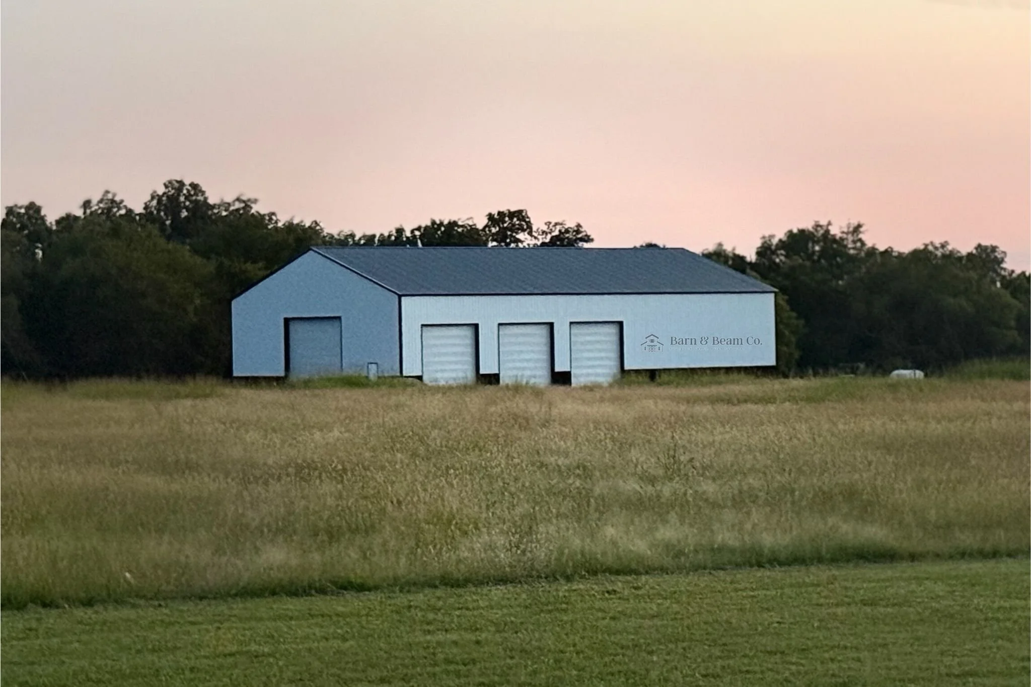 A large blue outbuilding with three garage doors and a smaller door, situated in a grassy field with trees in the background, during dusk.