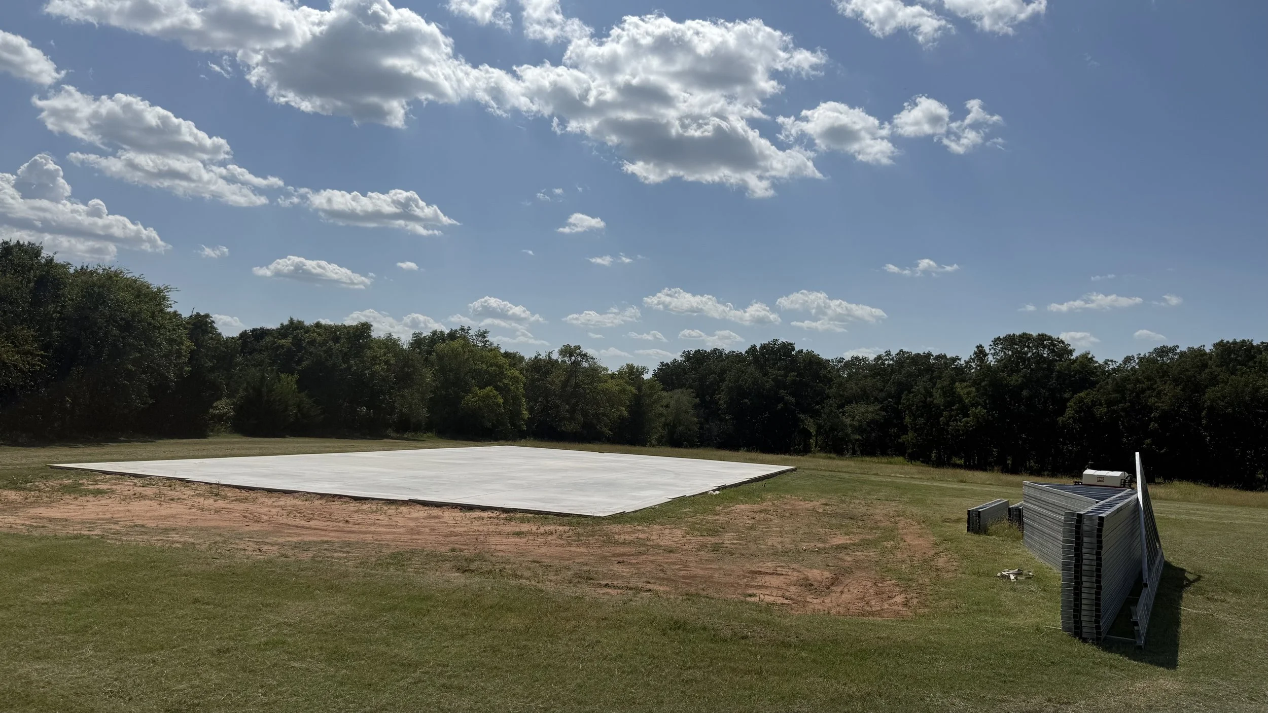 A large outdoor space with a white tarp or cover on the ground, surrounded by green grass and trees in the background. There are some black and silver metal frames or structures leaning on the right side of the image under a bright blue sky with scattered clouds.