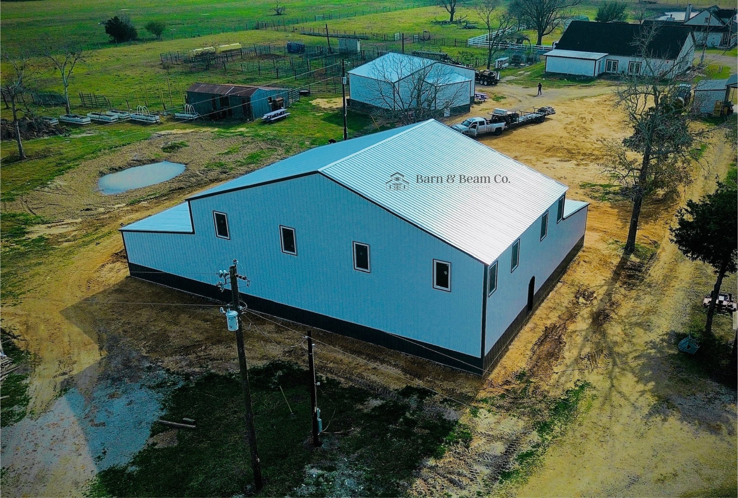 An aerial view of a large blue metal barn with a silver roof on a farm. Surrounding the barn are dirt paths, utility poles, trees, and other farm structures and equipment. The barn has a logo that reads "Barn & Beam Co." on the roof.