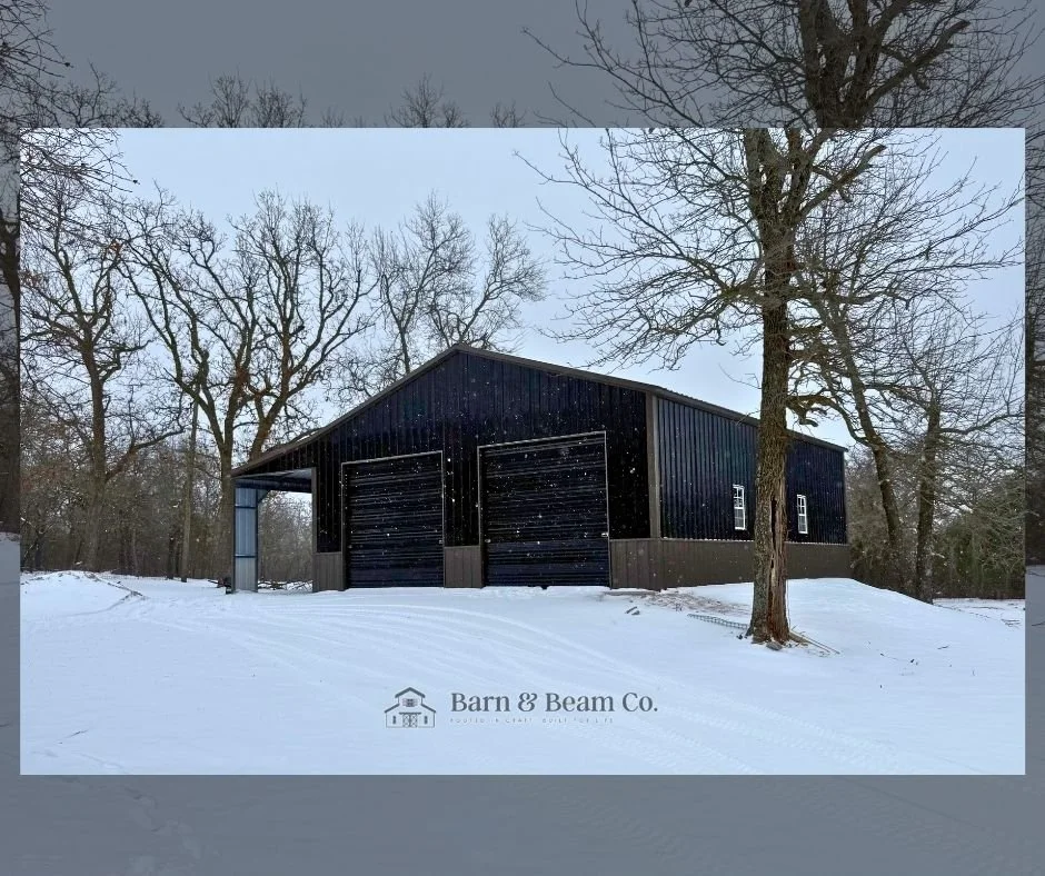 A large, black metal barn with two garage doors is situated on snow-covered ground, surrounded by leafless trees during winter. The image features a small logo reading 'Barn & Beam Co.' at the bottom.