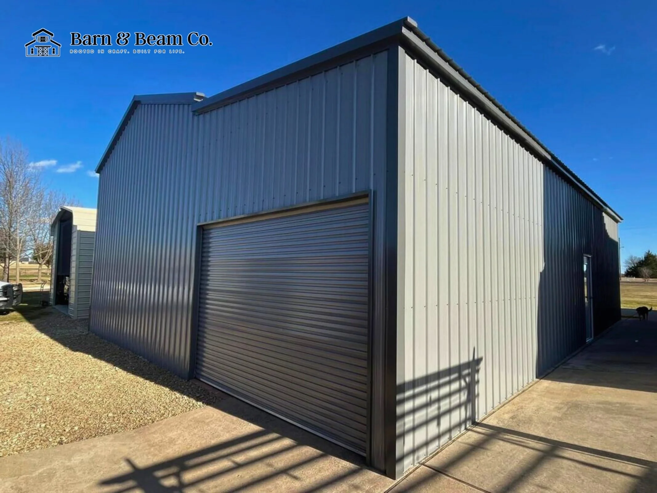 A large metal building with a roll-up garage door, sitting on a concrete and gravel surface under a bright blue sky.