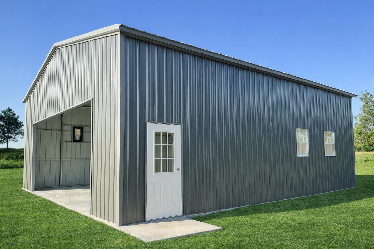 A metal barn with a white door, two small windows, and a large open section, set on a grassy field with trees and a clear blue sky in the background.