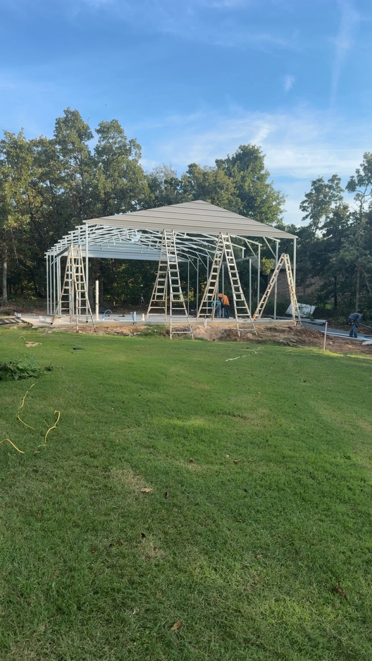 Construction workers building a metal framework for a large structure outdoors, with ladders leaning against it and a grassy area in the foreground.