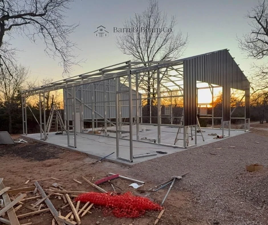 Construction site with a metal frame structure in progress, set on a concrete foundation, with a partially completed dark metal wall on one side, during sunset, surrounded by leafless trees and construction materials scattered on the ground.