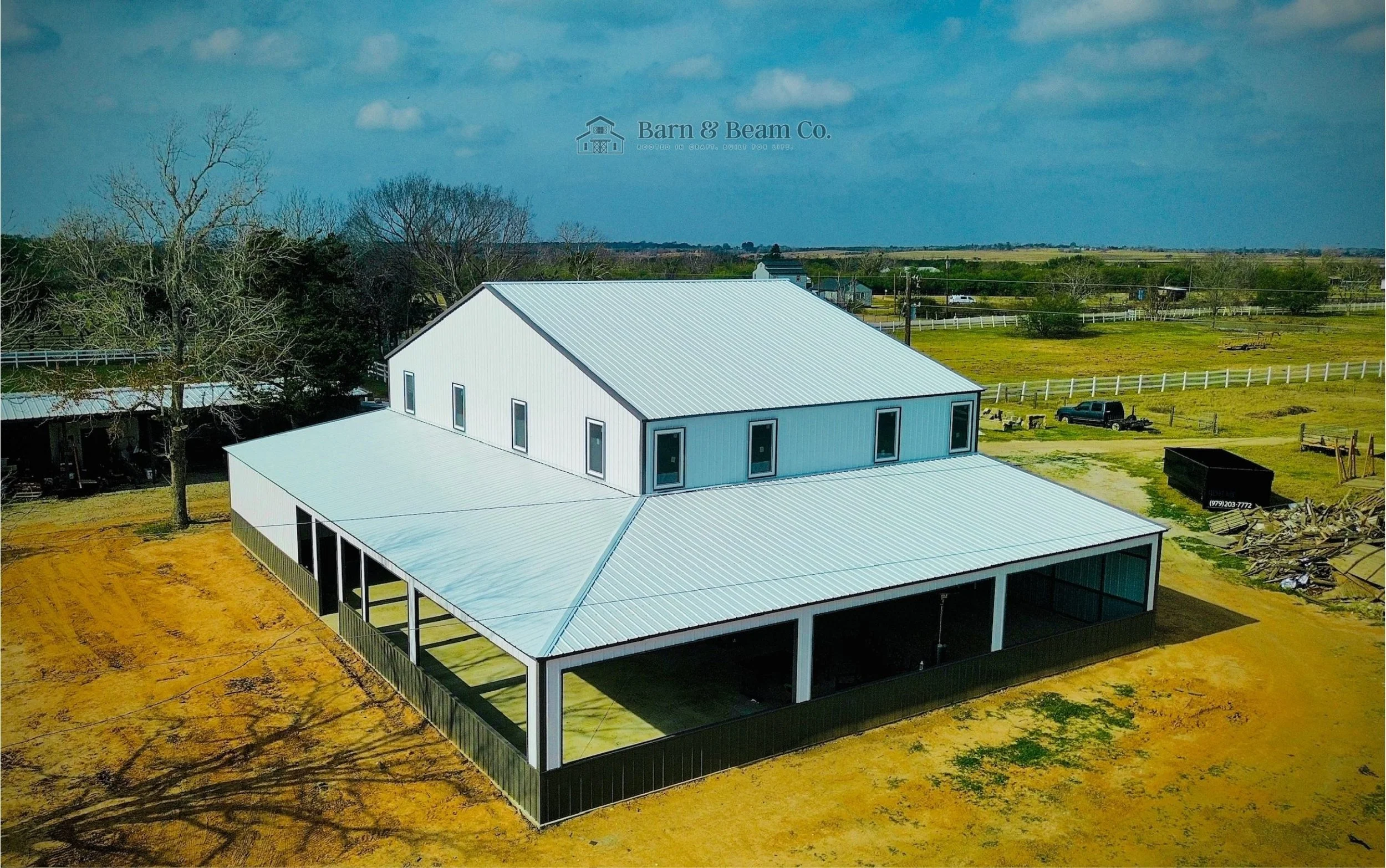 An aerial view of a large modern barn with a white metal roof in a rural area, surrounded by open fields, trees, and construction equipment.