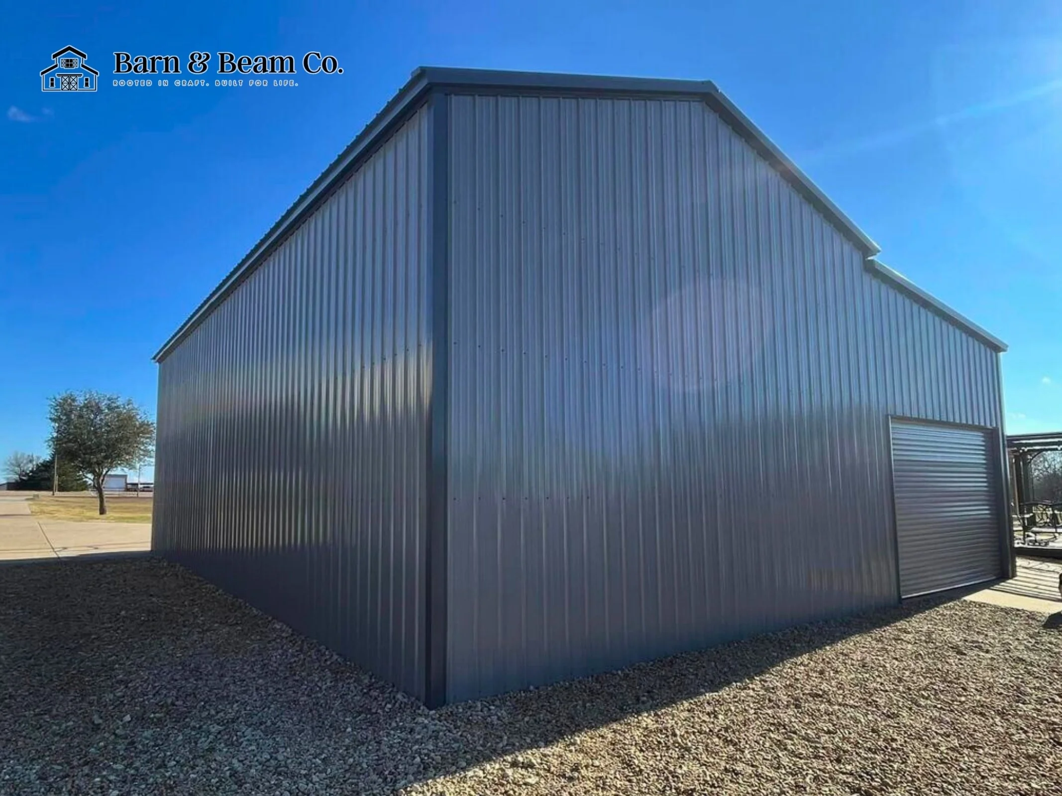 Large metal warehouse building with a roll-up door at the front, situated outdoors on gravel with a tree and open landscape in the background, under a clear blue sky.