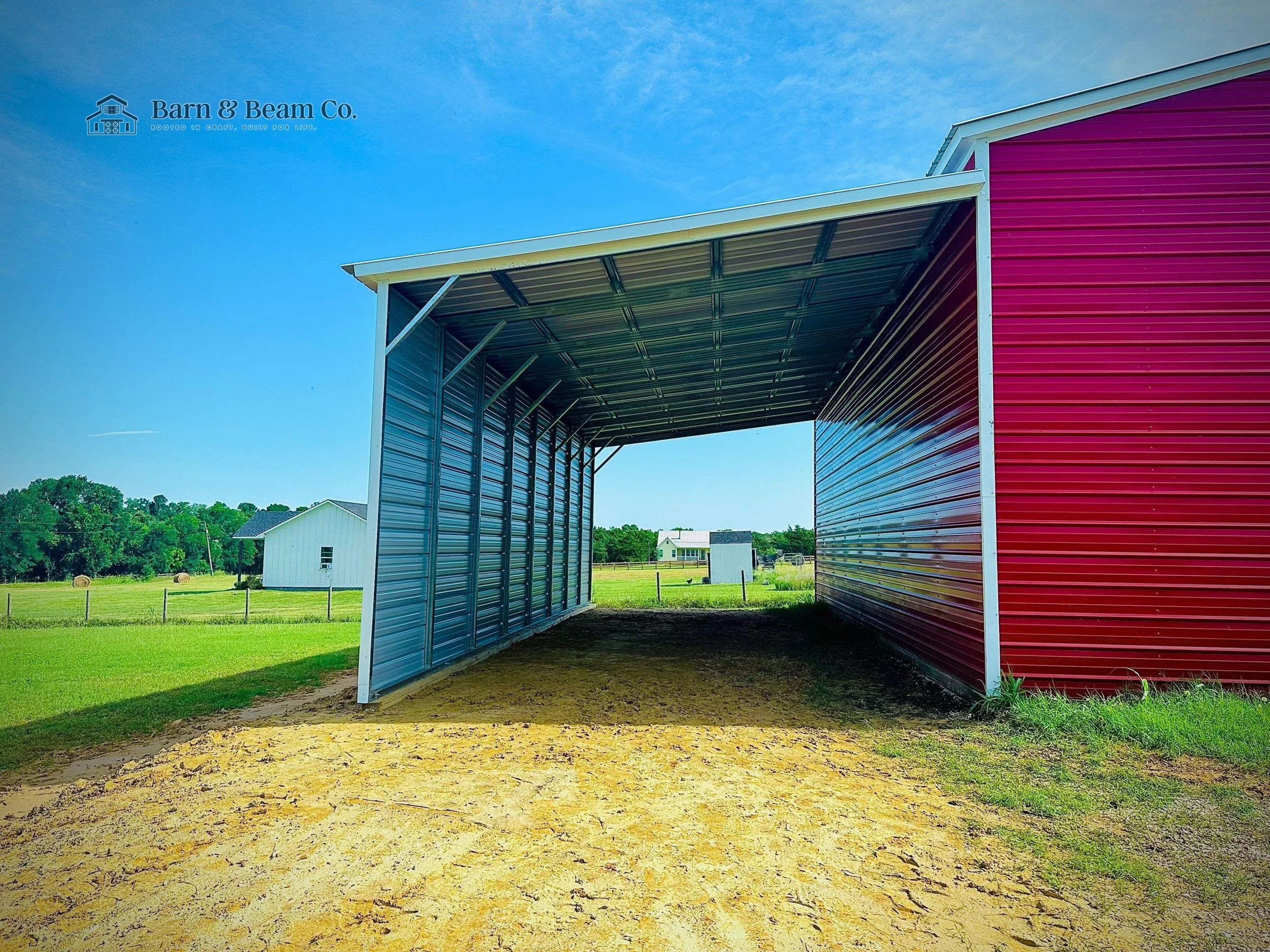 Metal red and blue barn on a sunny day with green fields and small white buildings in the background.