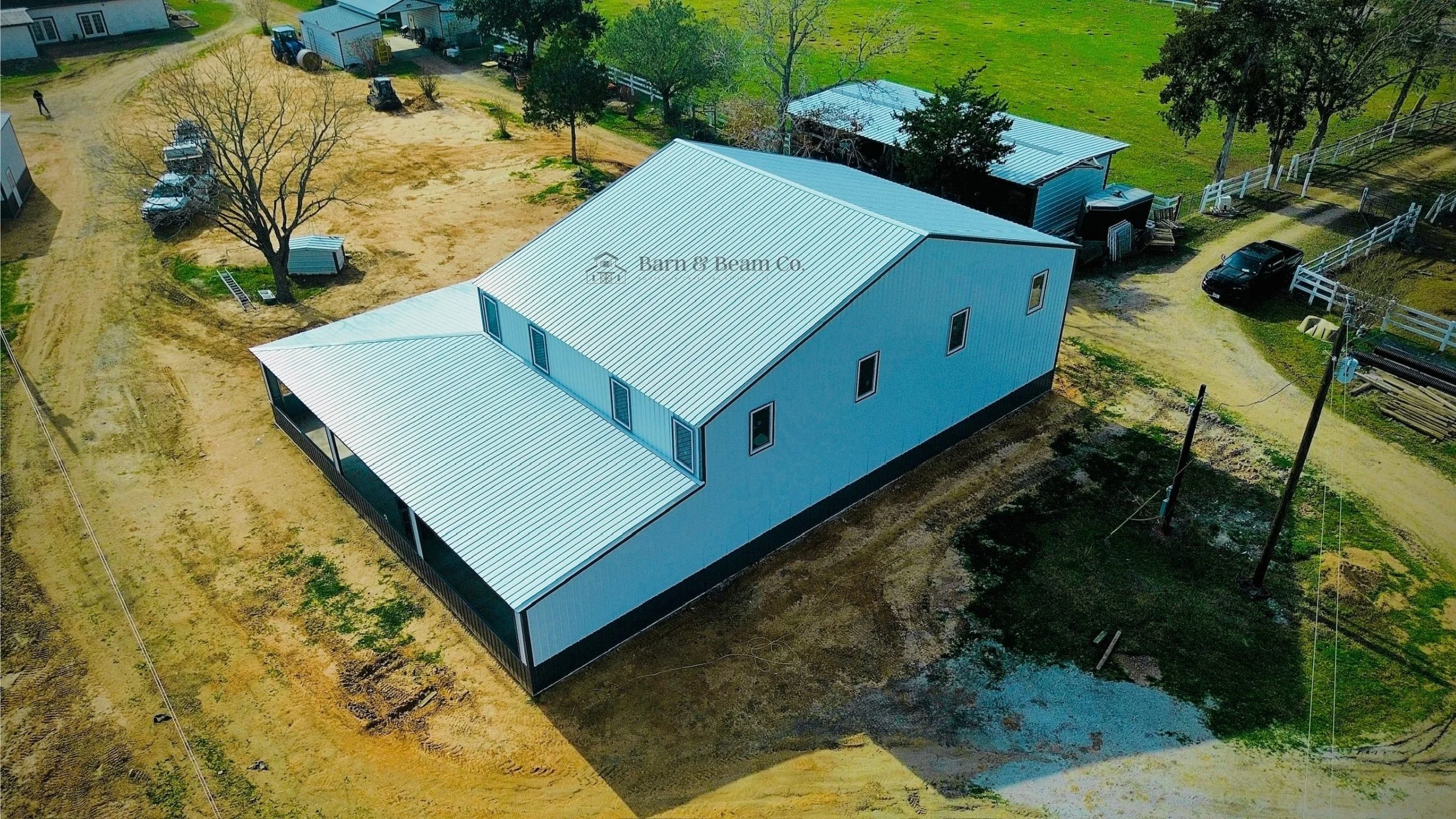 A metallic building under construction with a large sloped roof and several windows, situated on a cleared, partially muddy lot, surrounded by some trees and nearby smaller outbuildings on a farm or rural property.