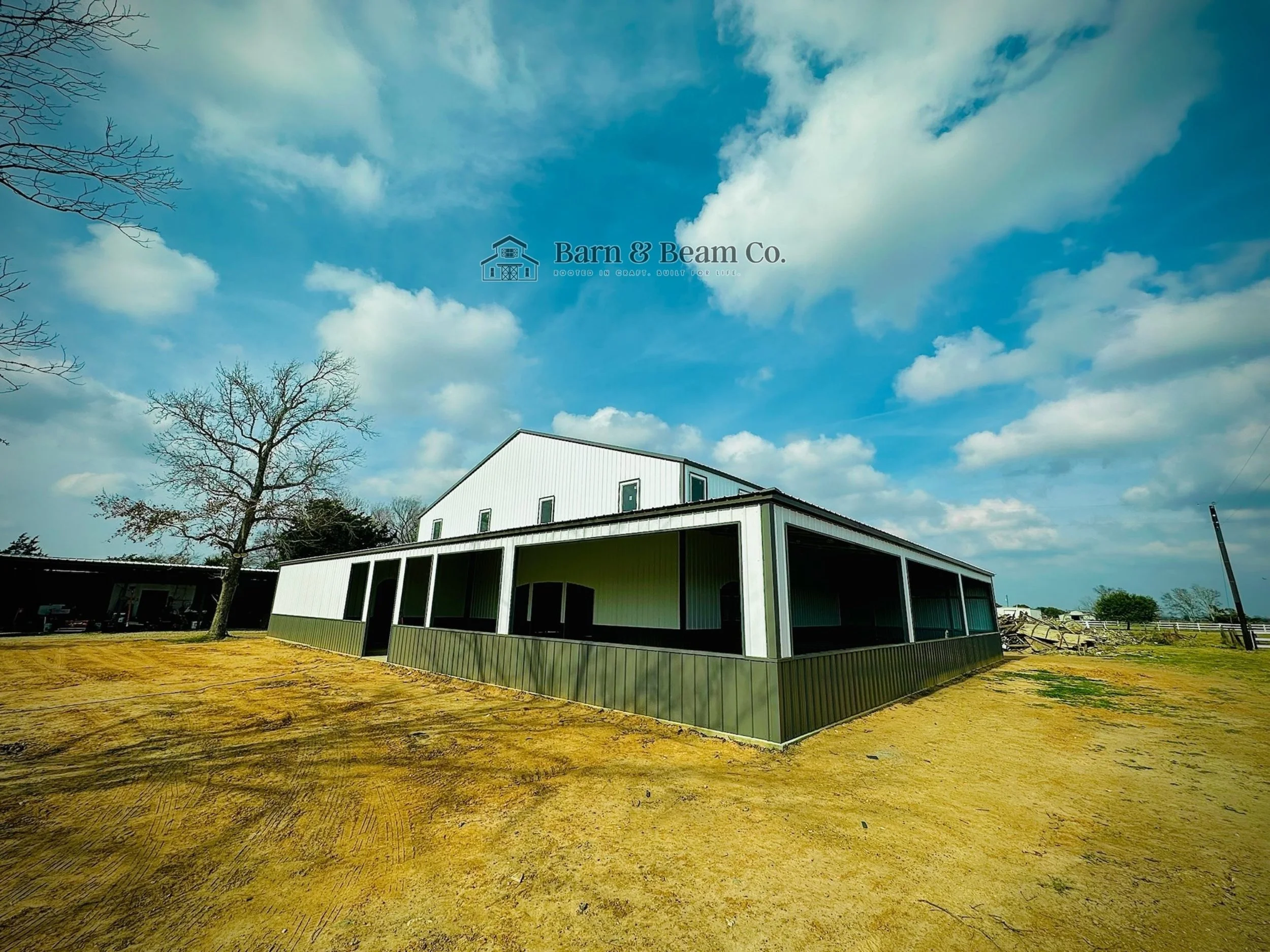 A large white metal barn with a screened porch in front, set on a dirt lot under a partly cloudy blue sky, with leafless trees nearby.