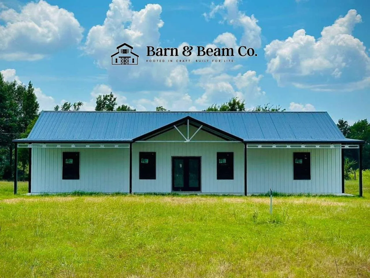 A white metal building with a black roof and trim, set in a grassy field with trees in the background under a partly cloudy sky. The building has four black doors, one in the center and three on either side, and a logo for Barn & Beam Co. with a barn