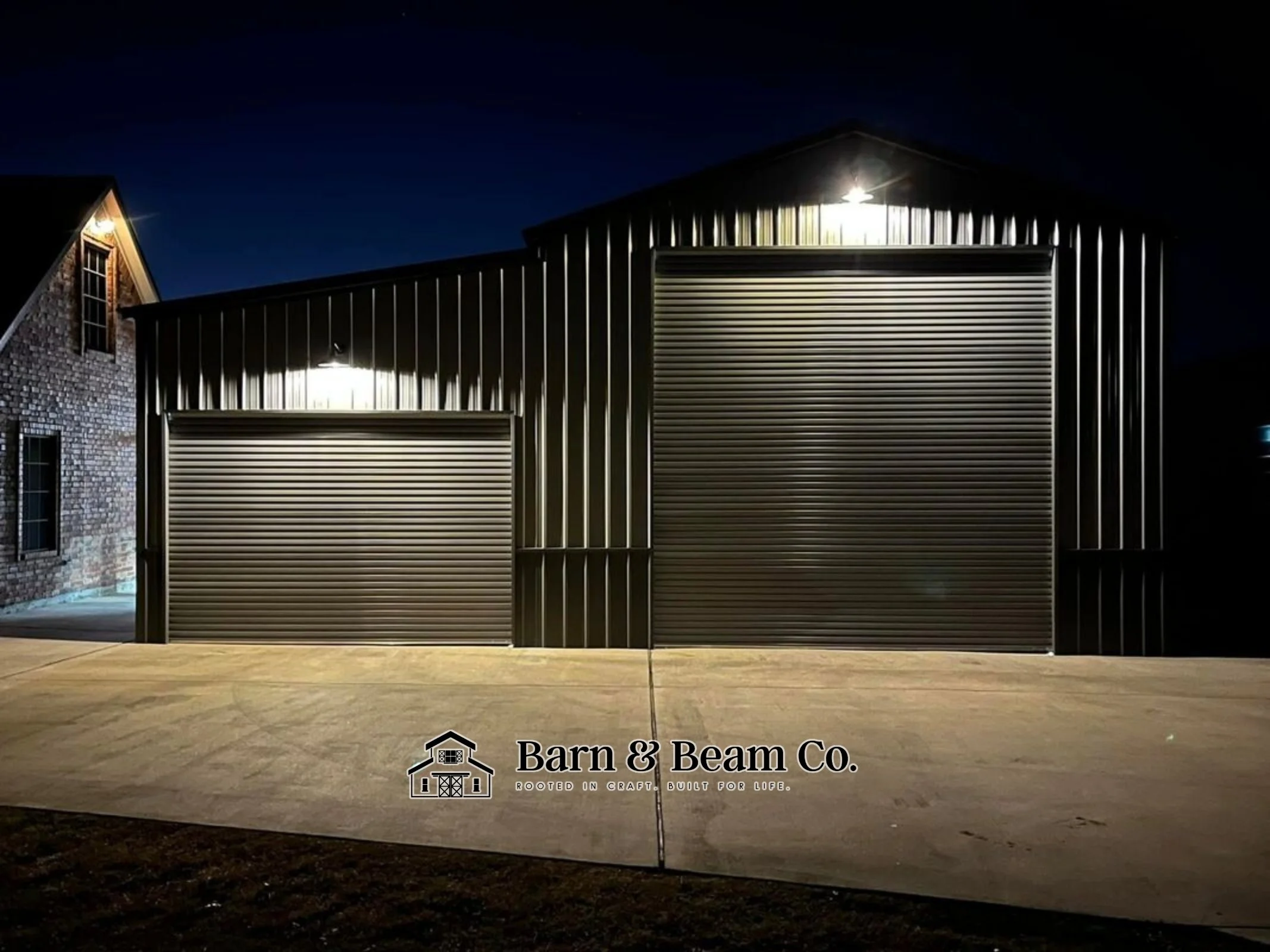 Nighttime exterior view of a metal barn with two large roll-up doors, illuminated by exterior lights, with a brick building to the left and a concrete driveway in front, featuring the logo and text "Barn & Beam Co. Rooted in craft. Built for life."