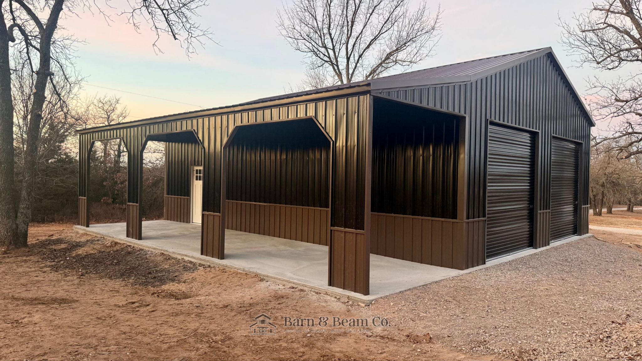 A black metal barn with a brown wooden lower section, two roll-up garage doors, and an open sheltered area with wooden posts, located in a rural setting with leafless trees and dirt ground.